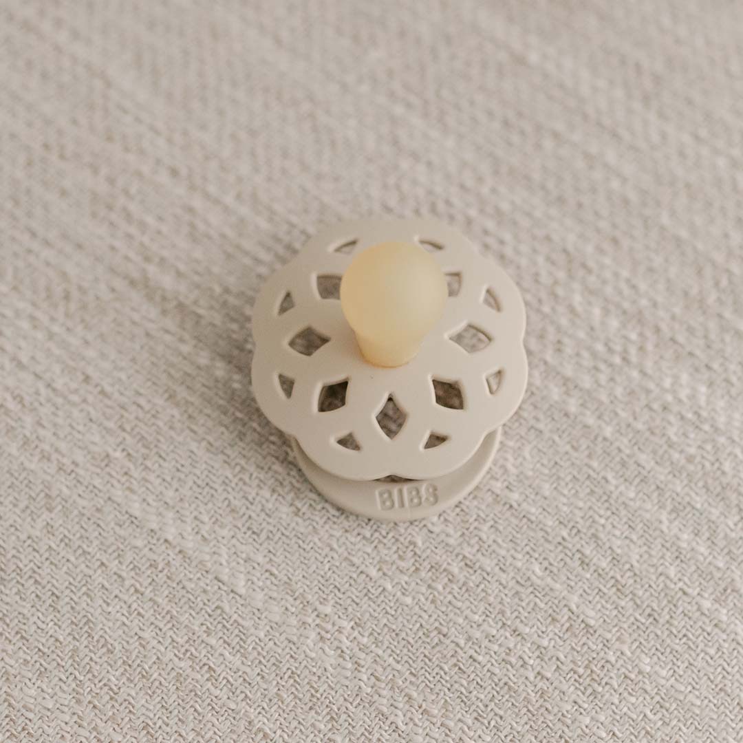 A vanilla pacifier with a decorative cut-out pattern rests on a textured fabric surface. Crafted from natural rubber, this Kristina pacifier from Denmark features a round nipple designed to help babies self-soothe. The brand name "BIBS" is elegantly displayed at the base.
