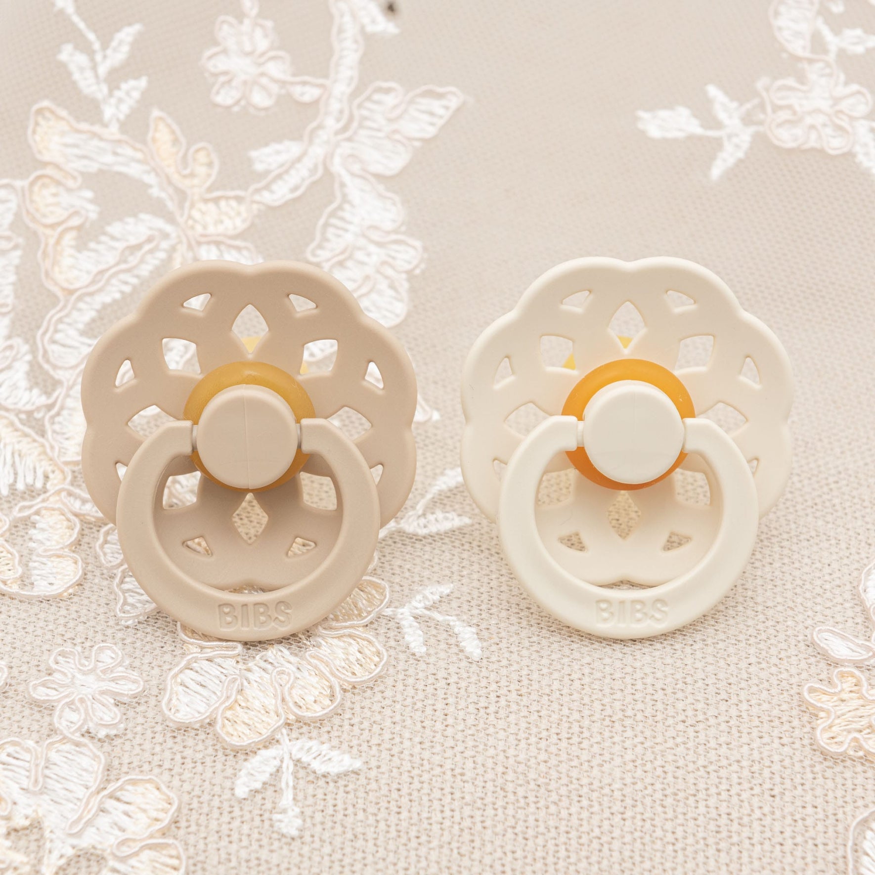 The Kristina Pacifier Set in Vanilla and Ivory features two decorative baby pacifiers crafted from natural rubber, beautifully displayed on an embroidered fabric background. These Danish designs, encouraging self-soothing for little ones, come with cut-out patterns, neutral colors, round handles, and have "BIBS" embossed at the bottom.