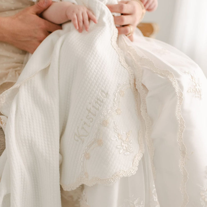 A close-up of a baby held by an adult, wrapped in the Kristina Personalized Blanket, which is adorned with light thread stitching of "Kristina" on quilted cotton featuring delicate lace and floral patterns.
