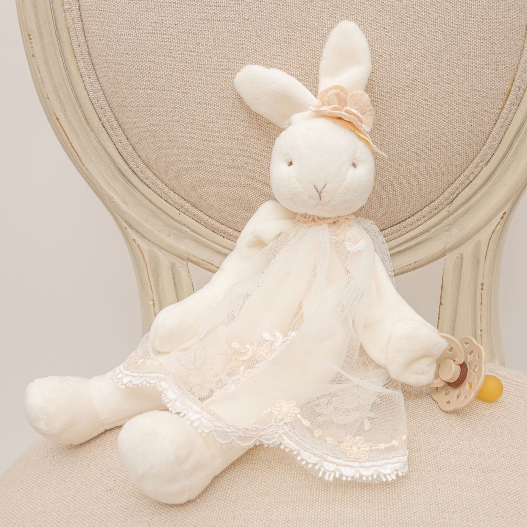 A Kristina Silly Bunny Buddy, dressed in a lace outfit and small hat, sits on a beige cushioned chair while holding a pacifier. The chair features a round back with a light rustic wood finish.