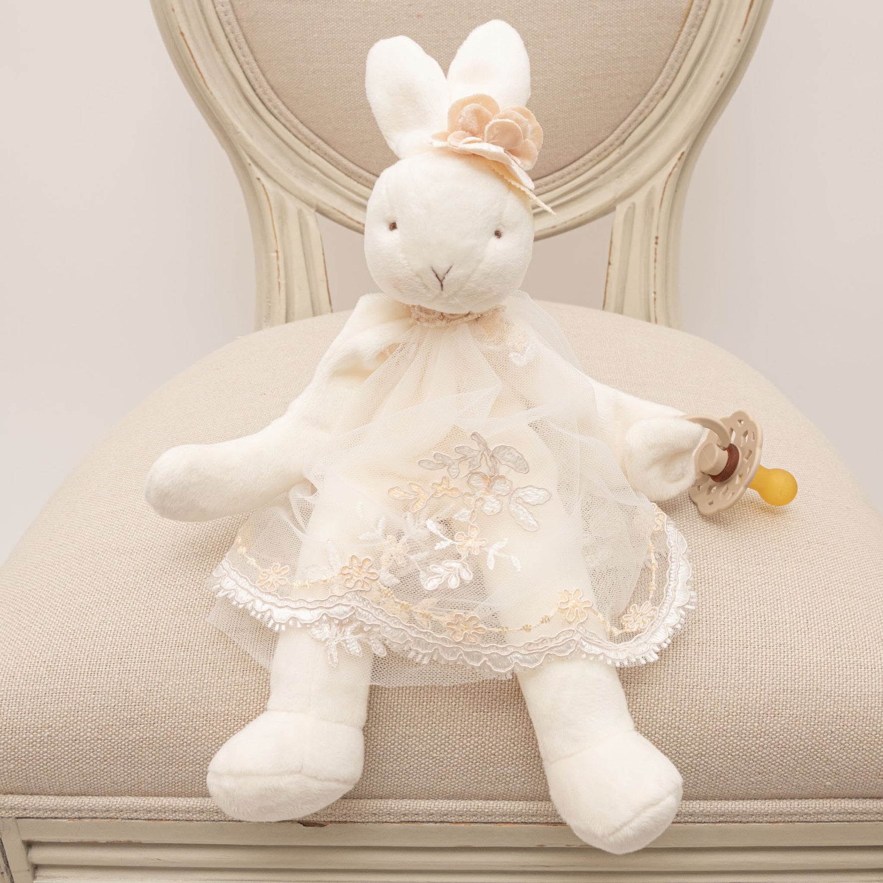 The Kristina Silly Bunny Buddy, a plush bunny, is seated on a beige upholstered chair. It wears a lace dress adorned with floral embroidery and holds a pacifier in one hand. The bunny also features a floral accessory on its head.