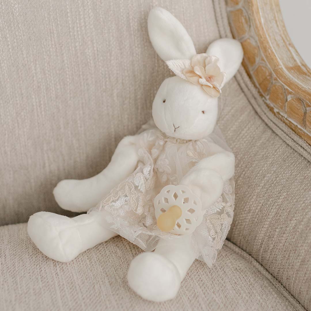 On a beige upholstered chair sits the Kristina Silly Bunny Buddy | Pacifier Holder, adorned in a lace dress and floral headband. It holds a small rattle and perfectly complements the soft, neutral color scheme—making it an ideal companion for any stroller adventure.