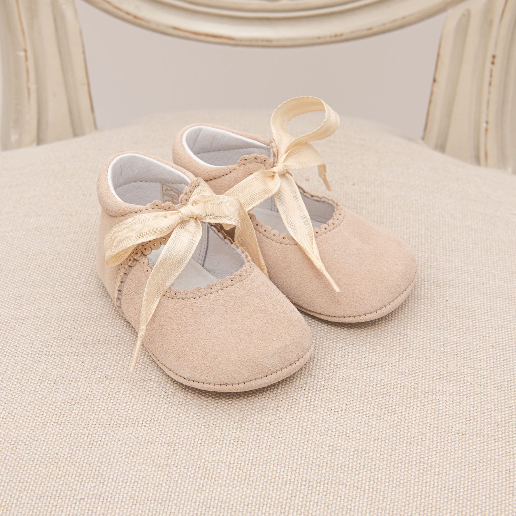 A pair of small, beige Kristina Tie Mary Janes with satin ribbon bows are placed on a cream-colored fabric surface. The handmade baby shoes feature scalloped edges and a soft, cushioned feel. A wooden chair with a beige finish is partially visible in the background.