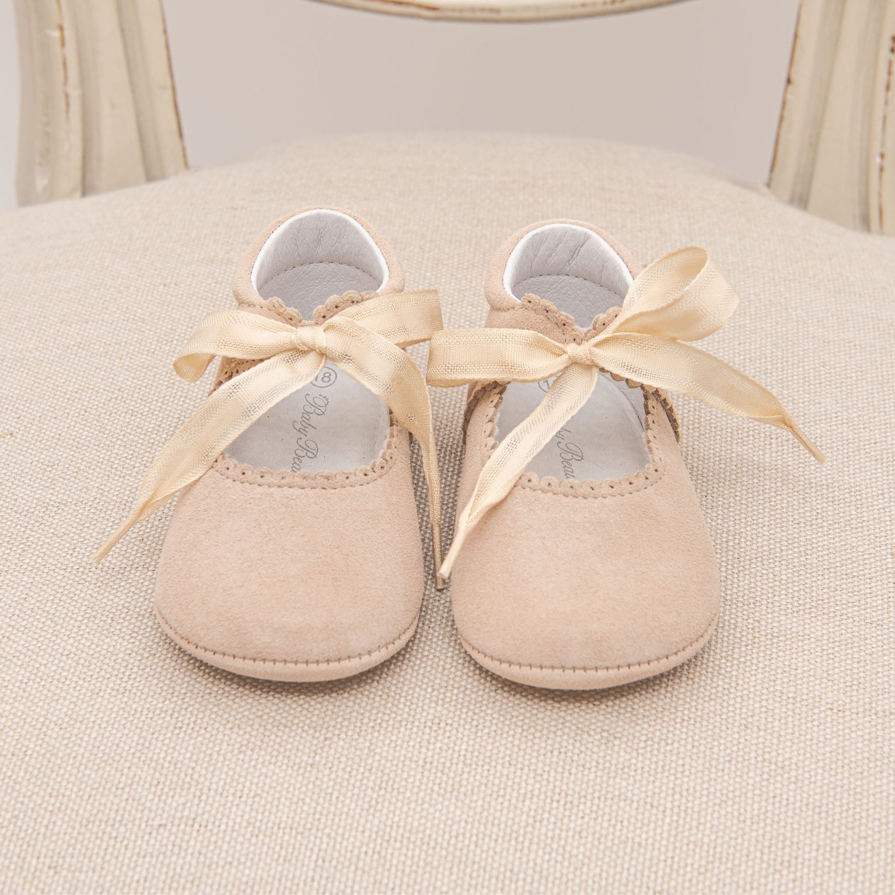 A pair of handmade Kristina Tie Mary Janes with satin ribbon ties, resting on a beige cushioned chair. The camel suede shoes have a scalloped edge design, evoking timeless elegance.