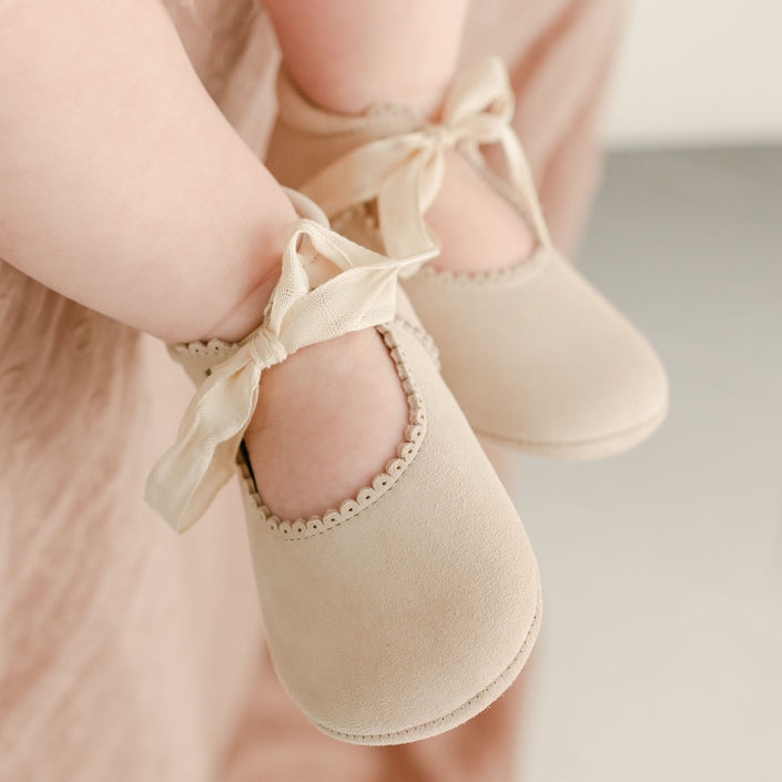 Close-up of a baby wearing handmade Kristina Tie Mary Janes with ribbon ties around the ankles. The shoes feature delicate scalloped trim around the opening. The baby is sitting, with a light-colored fabric visible in the background.