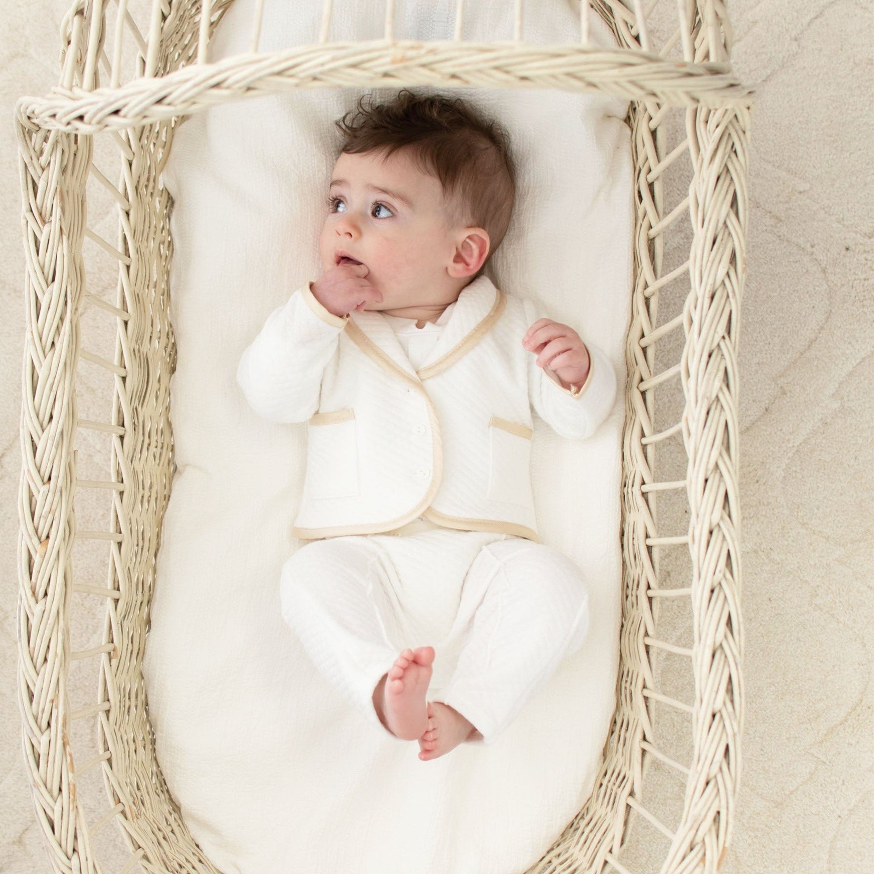 Baby laying in a cradle. He is wearing the Liam 3-Piece Suit made from a soft ivory quilted cotton and a champagne silk trim.