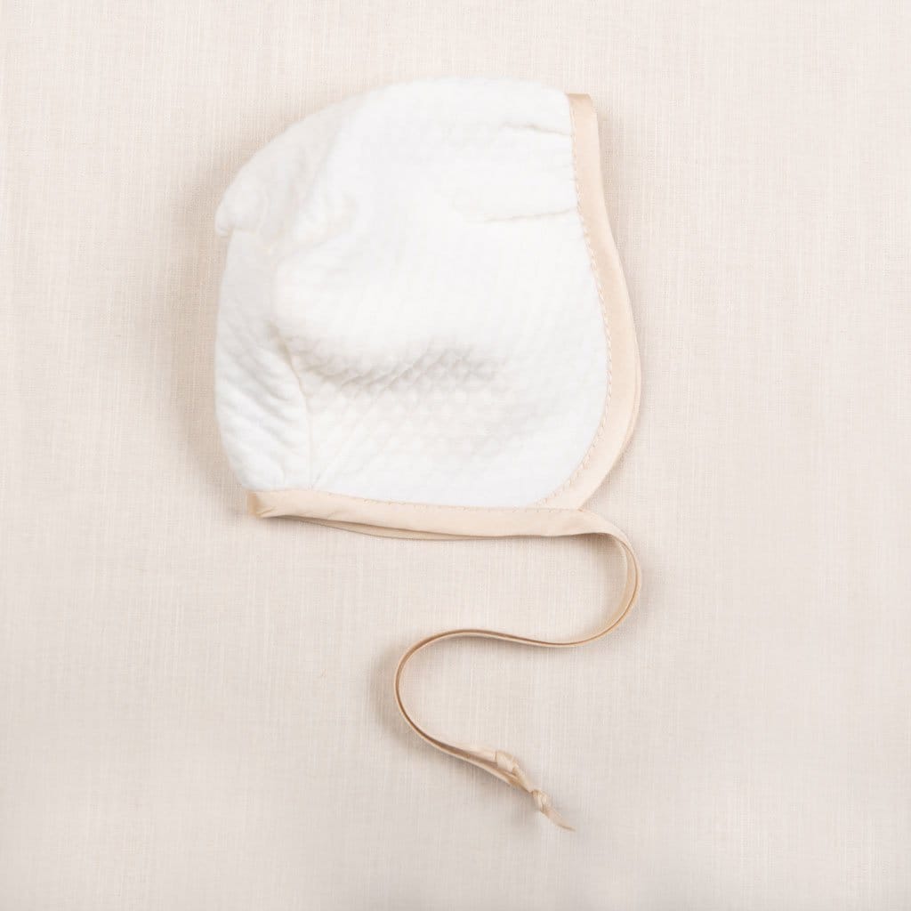 Flat lay photo of the Liam Quilted Newborn Bonnet made with soft quilted cotton in ivory and trimmed with a champagne colored silk