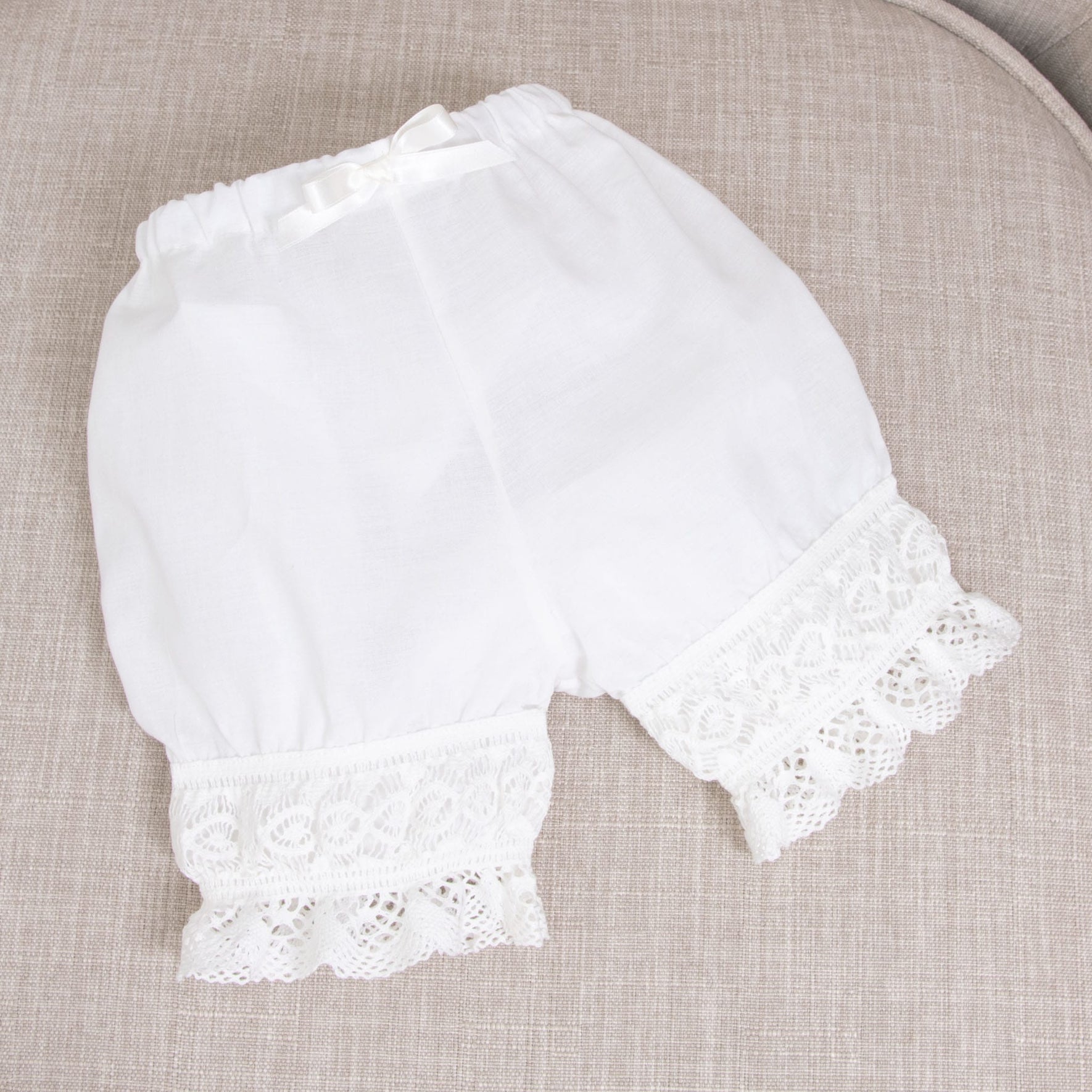 The Lily Bloomers, crafted from white linen and featuring a delicate lace trim, are displayed on a neutral, textured fabric background. A petite bow adorns the waistband, and these delightful pieces are meticulously handmade in the USA.
