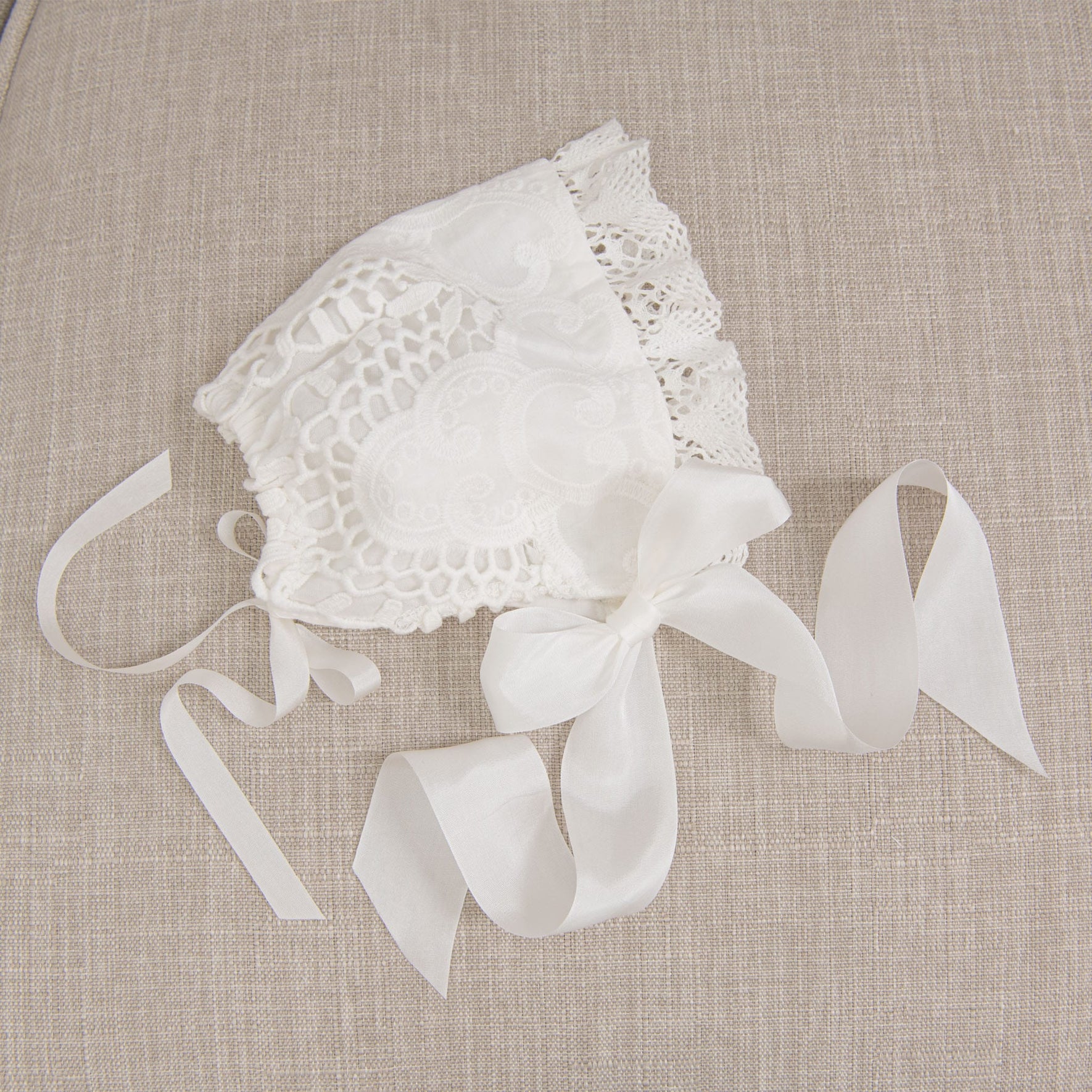 Flat lay of the Lily Christening Bonnet resting on a beige fabric surface, emphasizing the delicate light ivory cotton lace, intricate edge lace, and soft ivory silk ribbons.