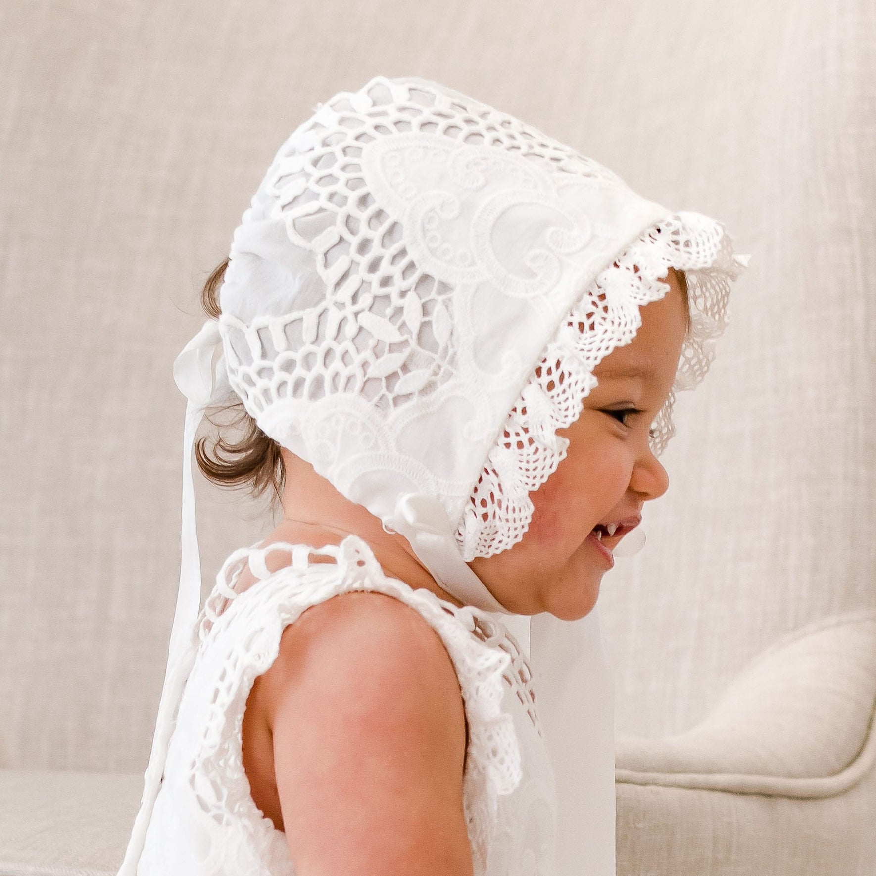 A baby girl in the Lily Christening Bonnet and matching dress looks away from the camera with a wide grin. The side profile highlights the bonnet’s delicate ivory cotton lace and intricate edge detailing.