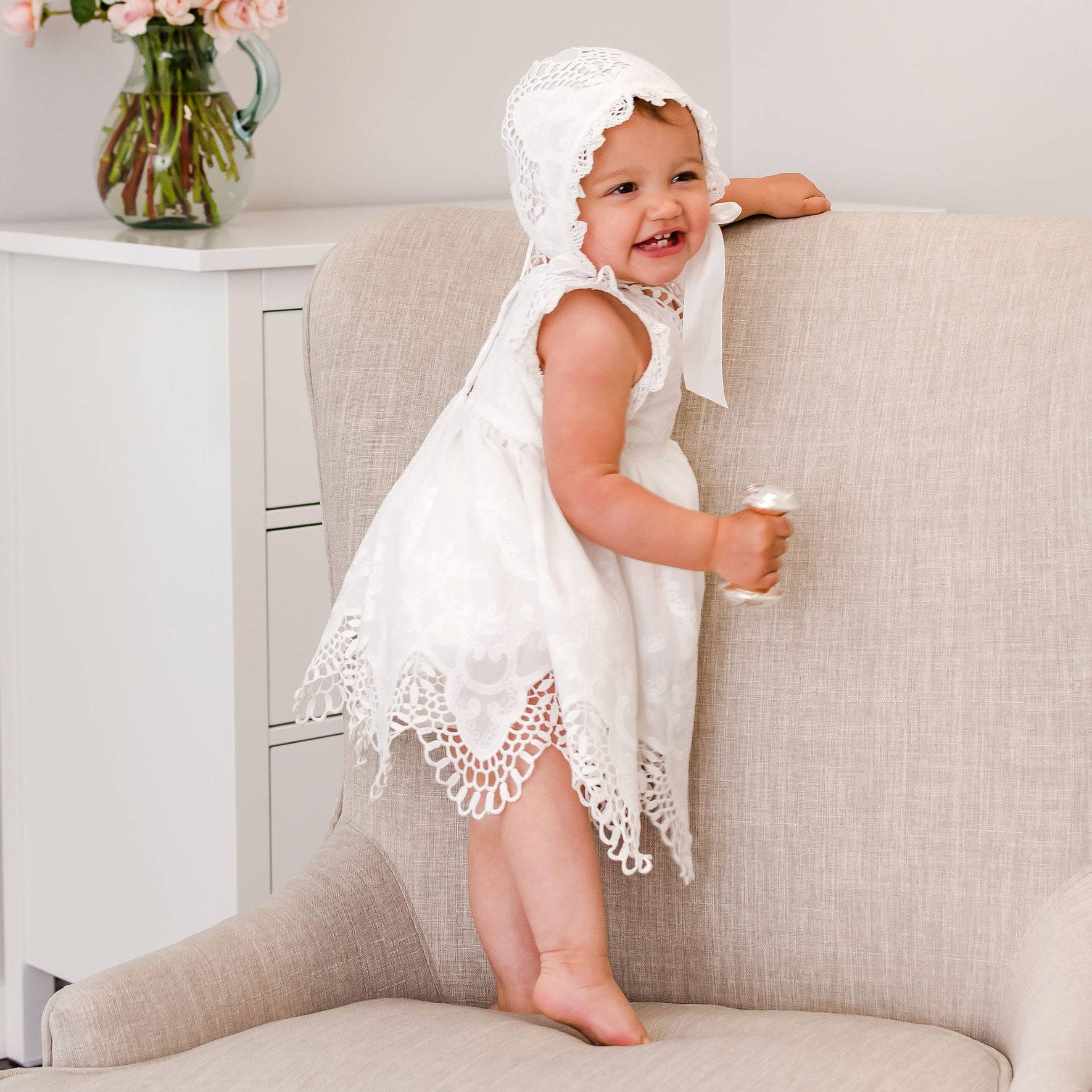 Baby on a chair and smiling as she wears a christening romper dress and holds a silver rattle