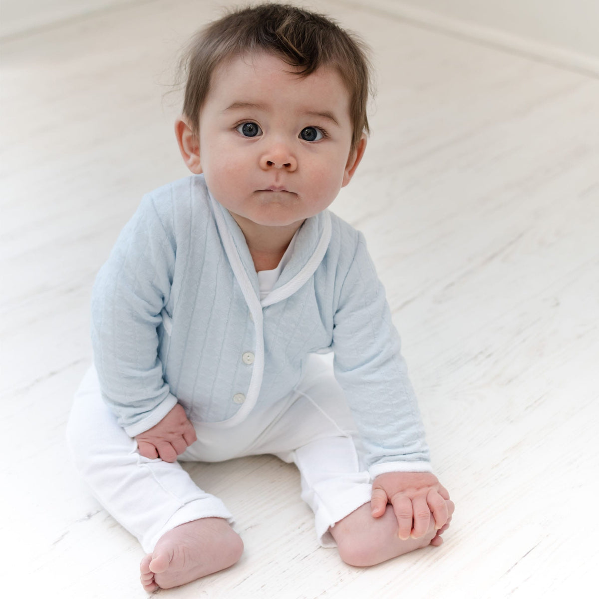 Baby boy sitting on the ground and wearing the Logan 3-Piece Suit, including the jacket, pants and onesie. Jacket made from blue textured cotton with a white French terry cotton trim.