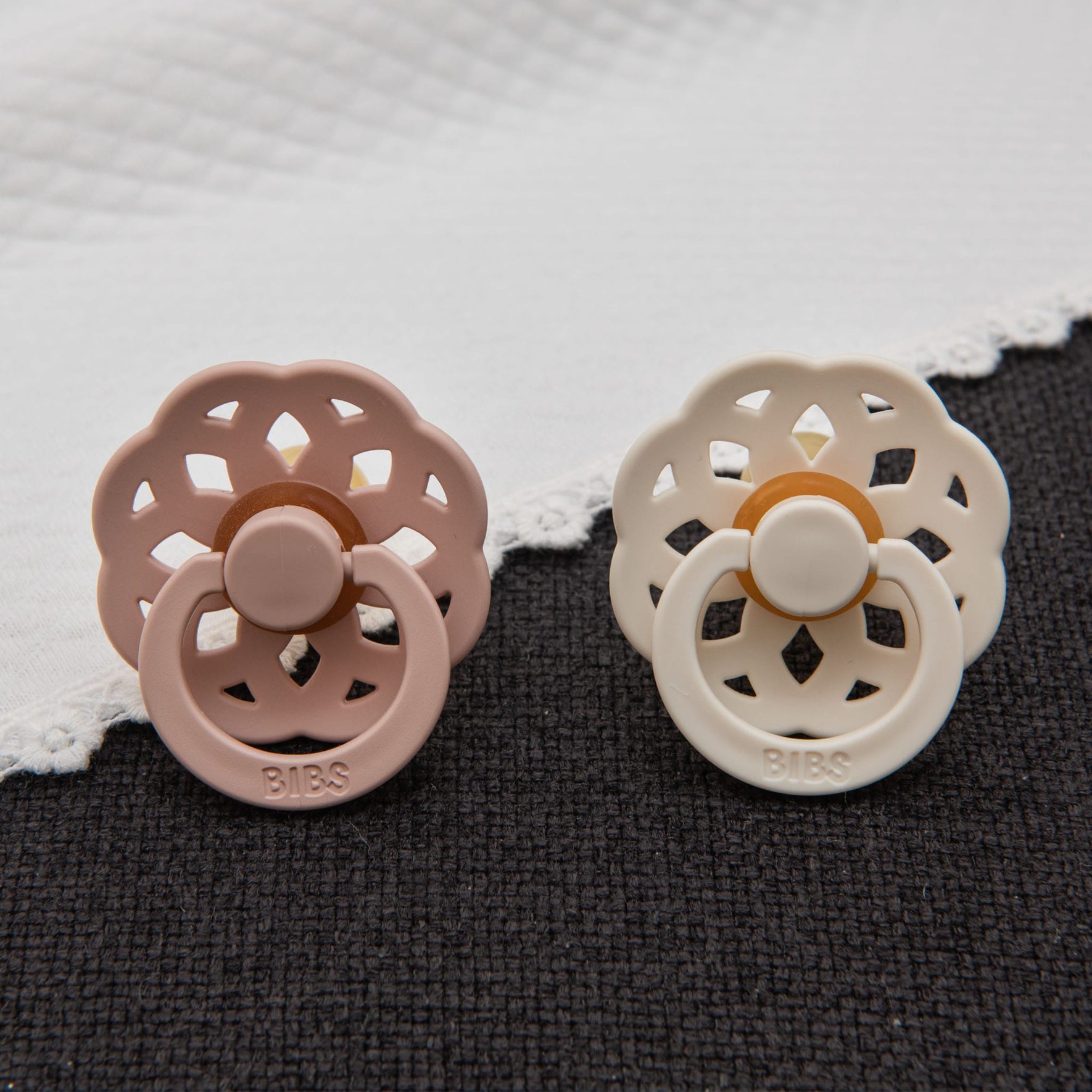 Close-up of blush pink and ivory pacifiers from the Lola Pacifier Set, placed on a dark fabric with a white blanket elegantly draped behind.