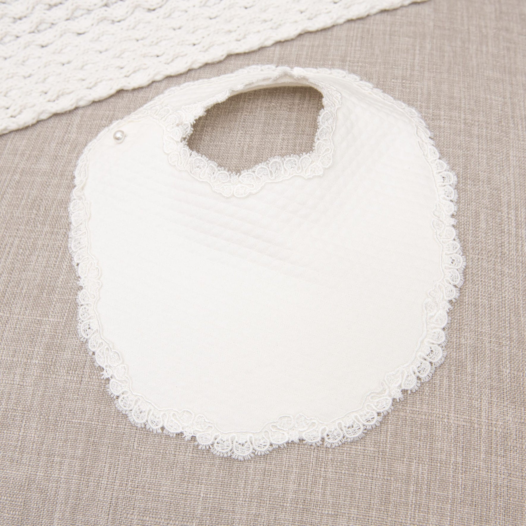 Displayed on a textured beige surface is a white baby bib with lace trim from the Madeline Accessory Bundle - Save 15%.