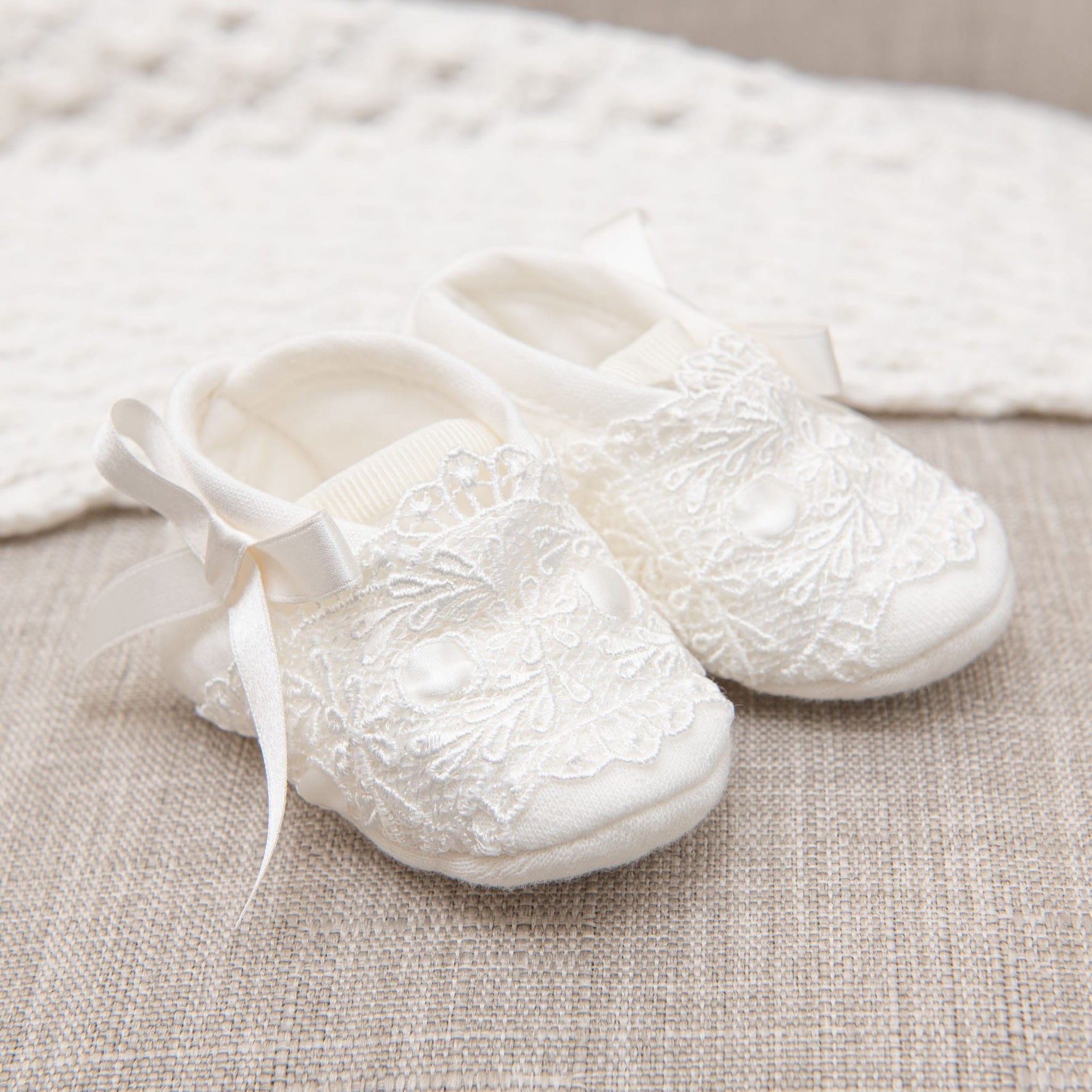 A pair of white baby shoes, adorned with delicate lace detailing and small bows, rests on a beige textured surface. Nearby, a knitted white blanket adds to the elegance, creating the Madeline Accessory Bundle - Save 15%, a charming newborn set perfect for welcoming little ones.