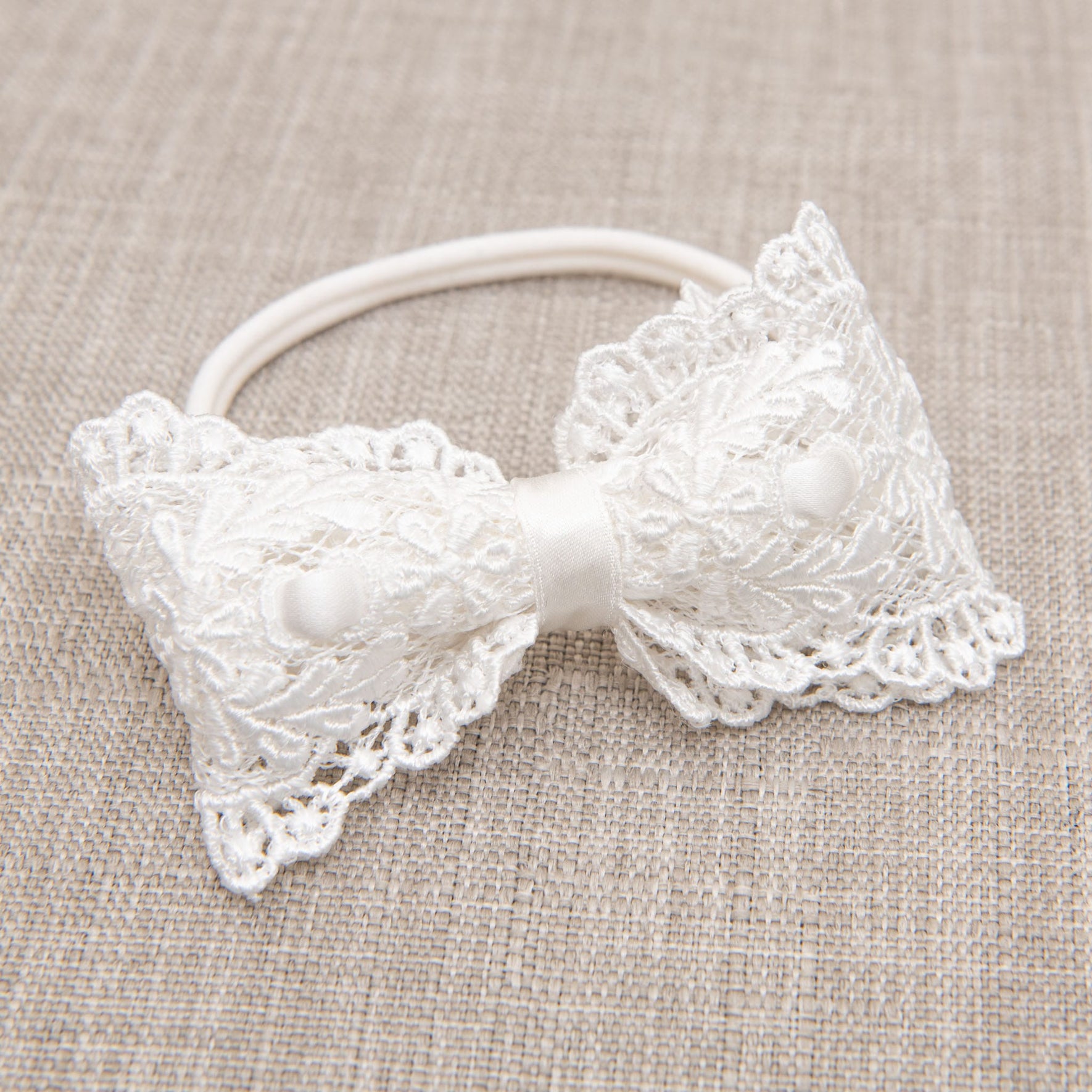 A Madeline Lace Bow Headband, featuring a white lace hair bow with intricate floral patterns and an ivory silk ribbon, is attached to an elastic band and placed on a beige textured fabric surface.