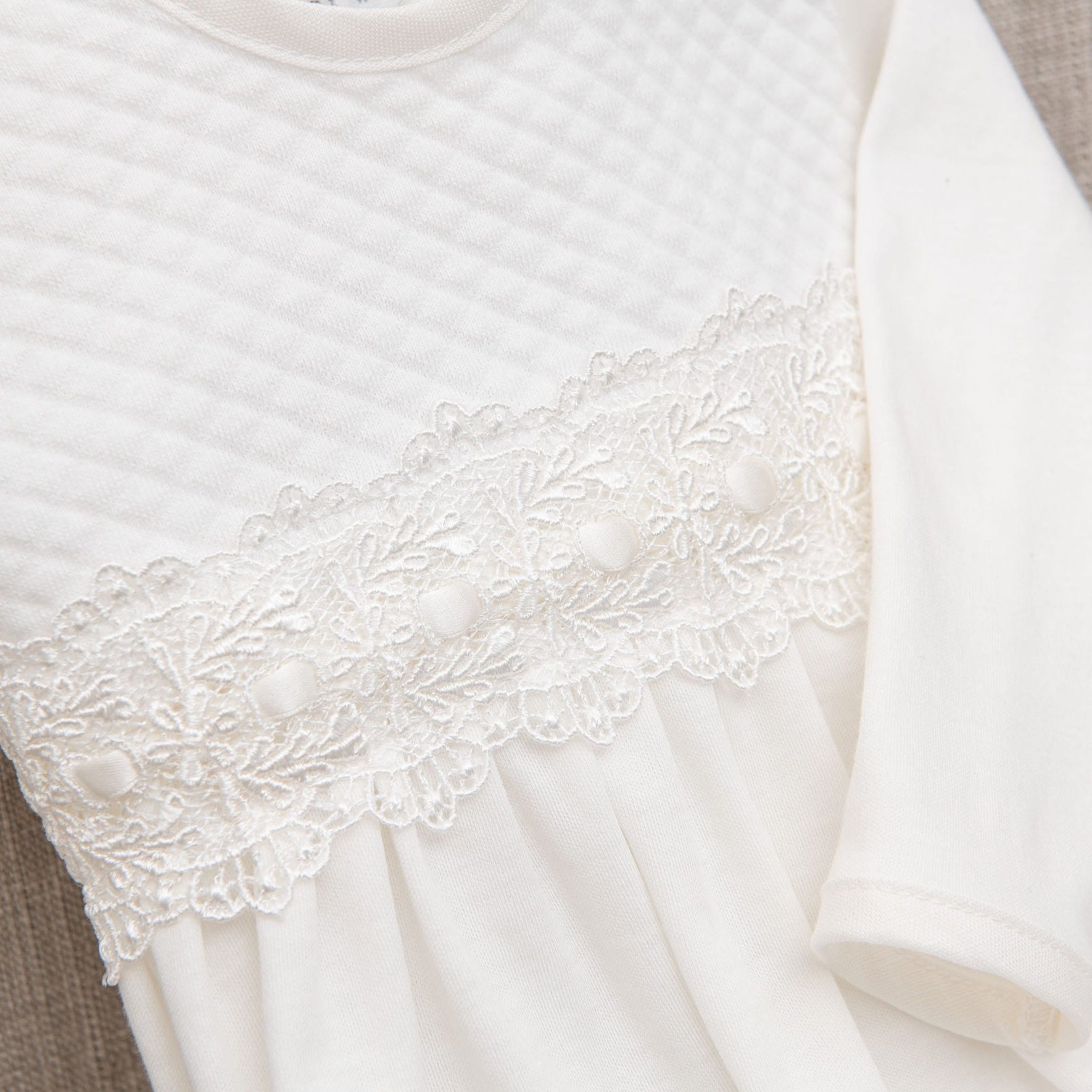 A close-up of the Madeline Newborn Gown crafted from soft pima cotton, featuring a quilted pattern on the upper section, delicate ivory lace trim with floral design and small fabric-covered buttons in the middle, and a pleated lower section. The sleeve on the right side is plain white.