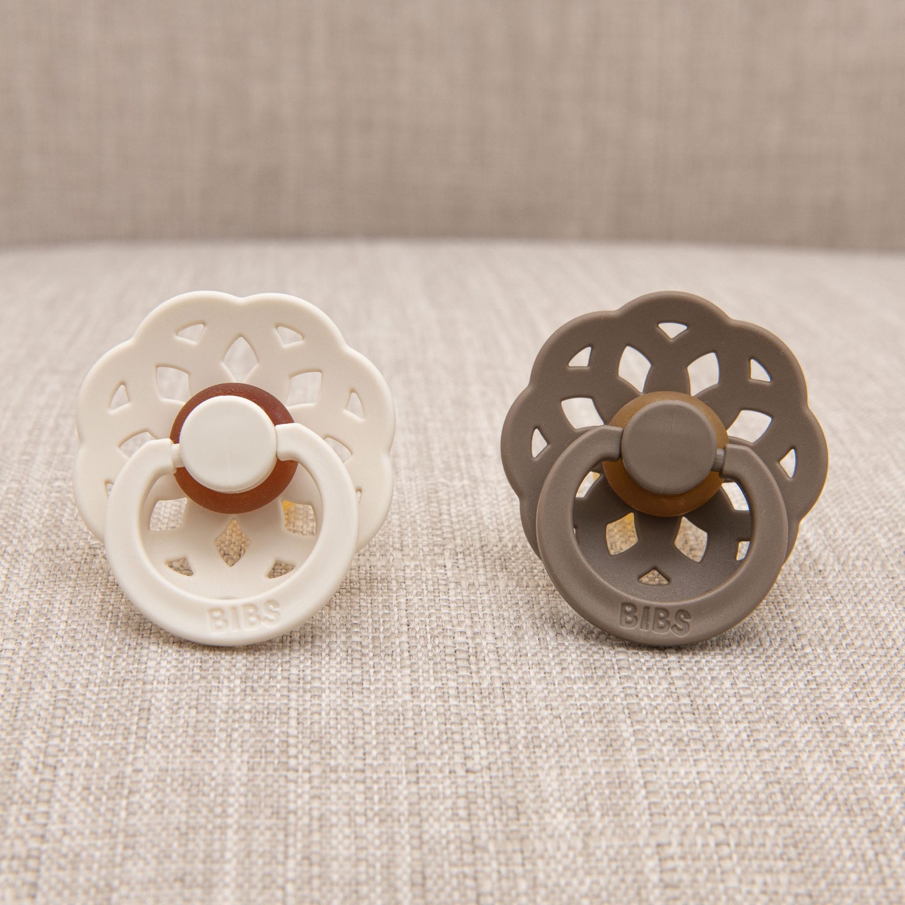Positioned side by side on a textured beige surface are two pacifiers. The pacifier on the left is ivory, while its counterpart on the right is dark oak. Both are meticulously crafted from BIBS natural rubber to aid in self-soothing. They feature scalloped edges and a circular center with "BIBS" elegantly embossed on the front—a testament to Danish craftsmanship. This set is part of the Madeline Pacifier collection.