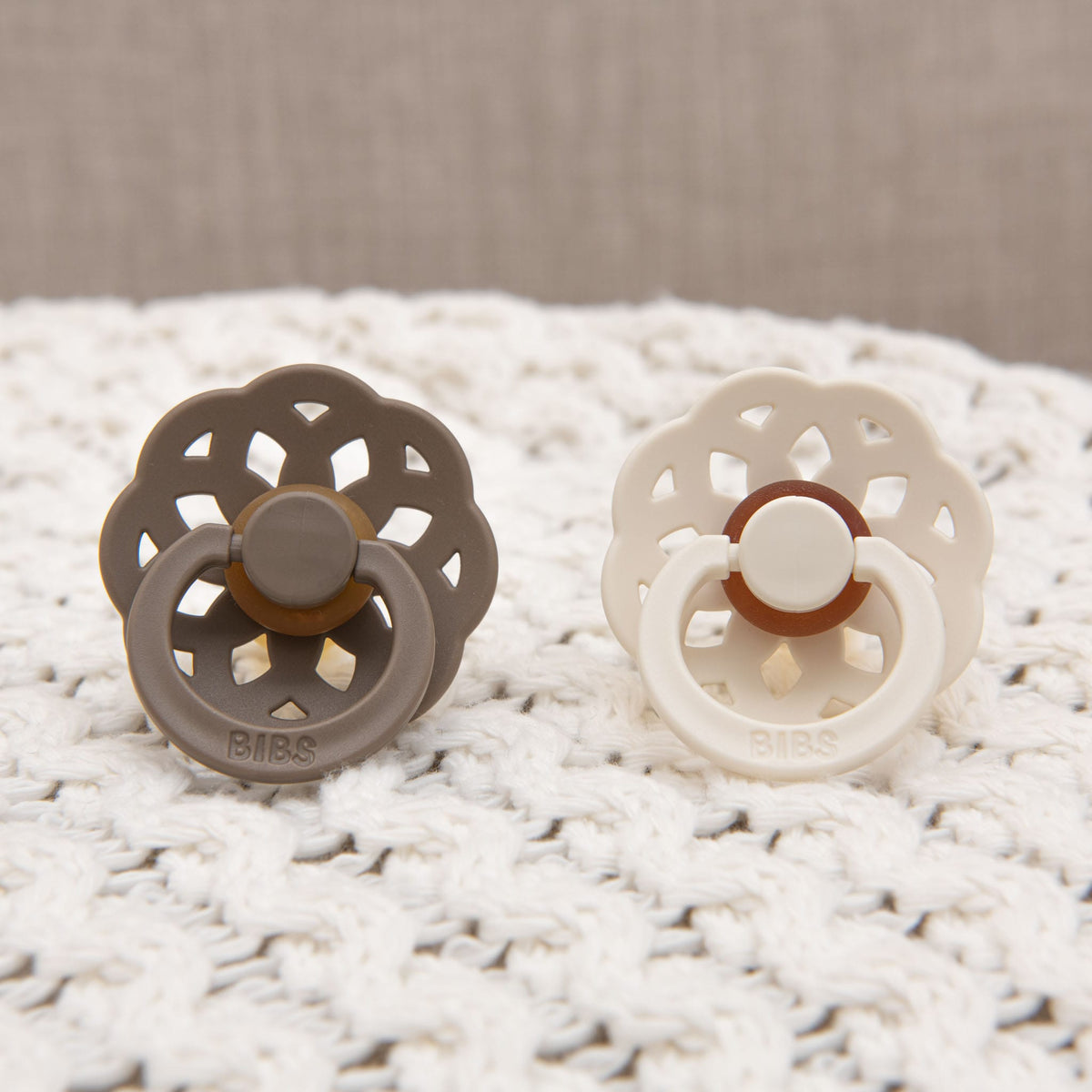 Two Madeline pacifiers with scalloped edges rest on a textured white surface. Crafted in Denmark, one is ivory and the other dark oak, both featuring a ring design with the brand name "BIBS" embossed on the front. These BIBS natural rubber baby pacifiers are designed to help babies self-soothe.