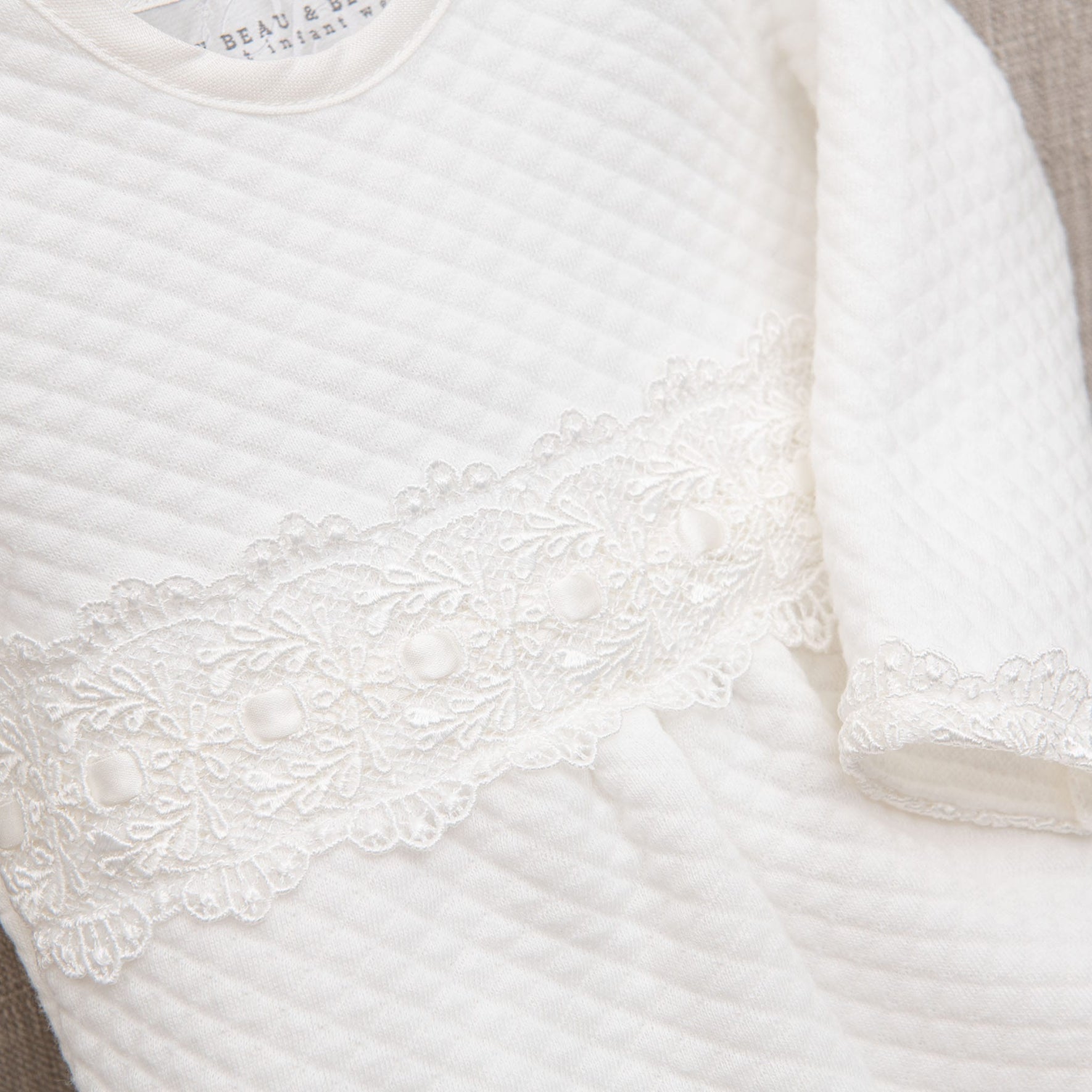 Close-up image of a white textured fabric garment with lace detailing near the waist. The lace features a floral pattern with small circular accents. The Madeline Quilted Newborn Romper, handmade in USA, has long sleeves and a round neckline.