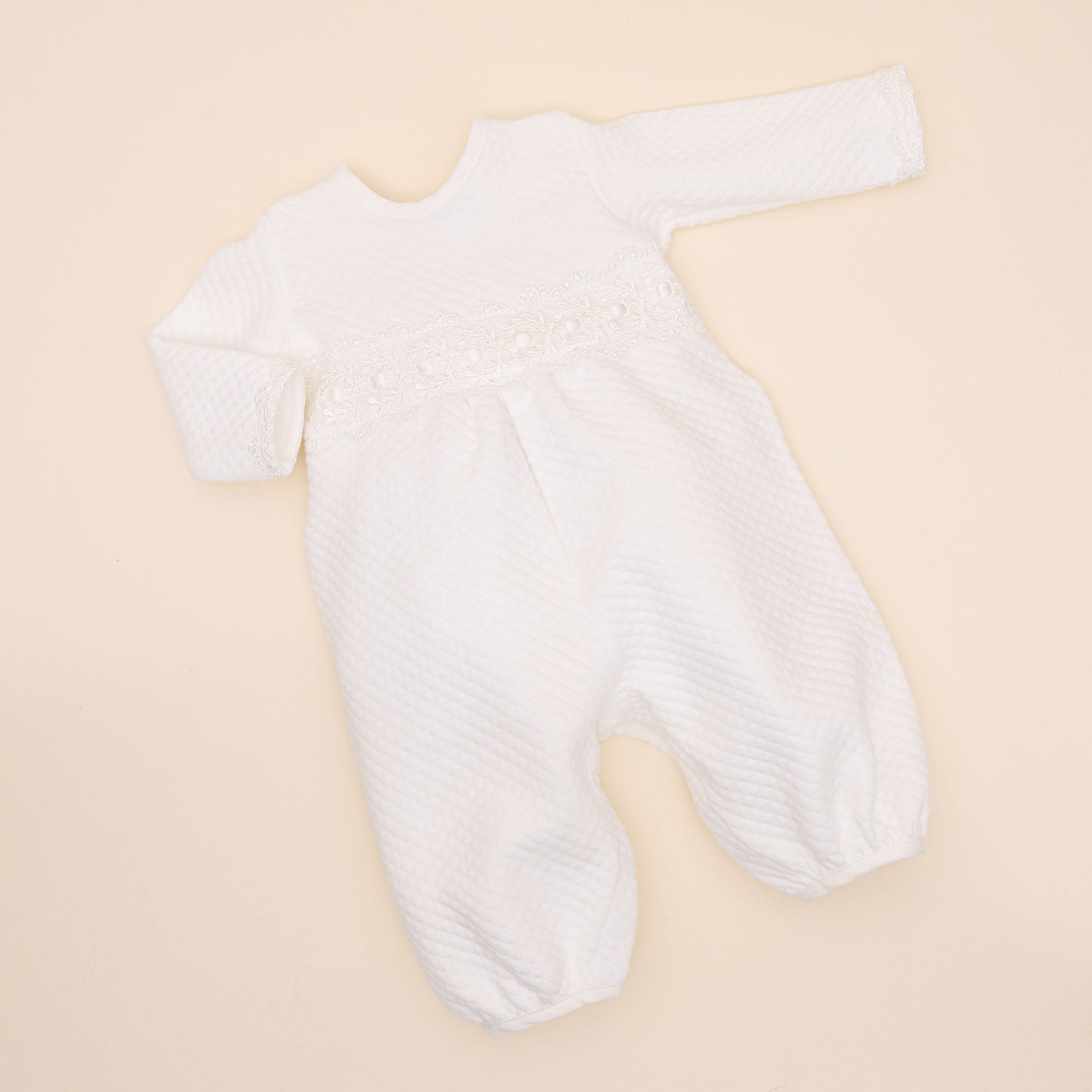 A white, long-sleeved Madeline Quilted Newborn Romper is laid out flat on a beige background. The onesie, handmade in the USA, has a textured pattern and features lace detailing across the chest area. The garment has a round neckline and closed feet.