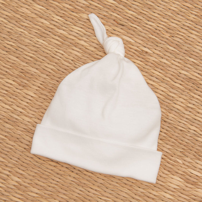 A Mason Knot Cap, made from soft pima cotton in white, rests on a woven wicker surface, making it perfect for a newborn christening.