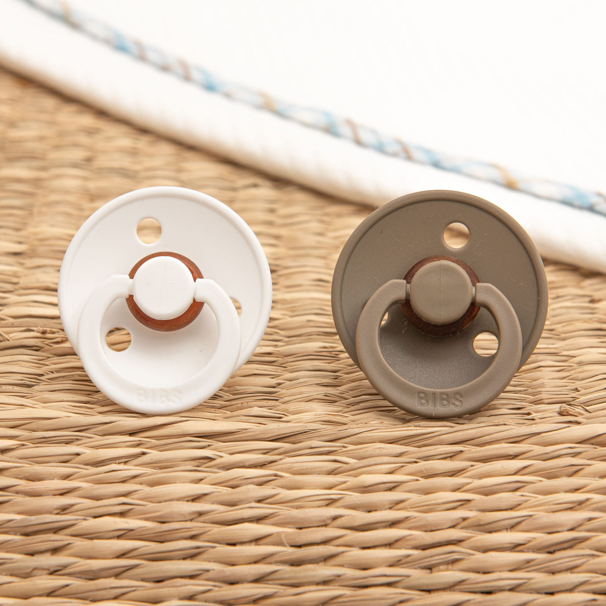 The Mason Pacifier Set in White and Dark Oak rests on a woven straw surface. Each pacifier, crafted from natural rubber for baby self-soothing, features a circular shield with two holes, a rounded nipple, and "BIBS" proudly engraved on them to signify their Denmark manufacturing.