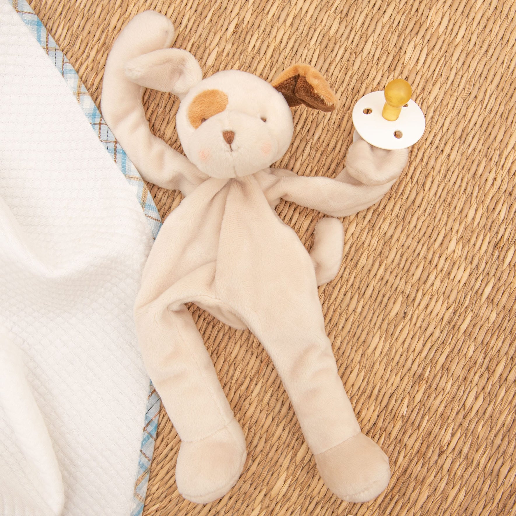 A Mason Puppy Buddy | Pacifier Holder, crafted from soft beige velour in the shape of a dog with floppy ears, rests on a woven surface. Featuring a stitched face and holding a yellow pacifier, it makes an ideal Christening gift. It is positioned next to a white blanket with partly visible plaid edges.