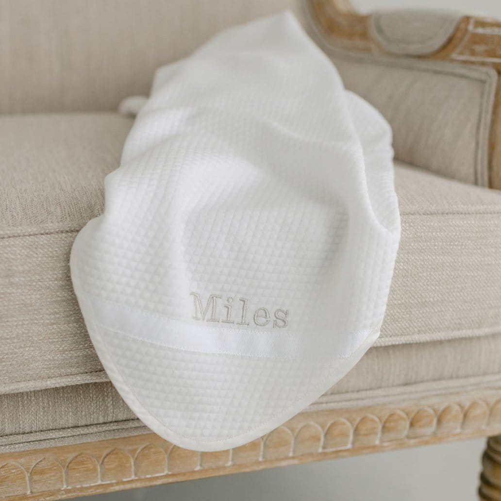 Flat lay photo of the Miles Personalized Blanket on a chair. It is made with quilted cotton and a ribbon detail on the corner. The name "Miles" is embroidered on the corner.