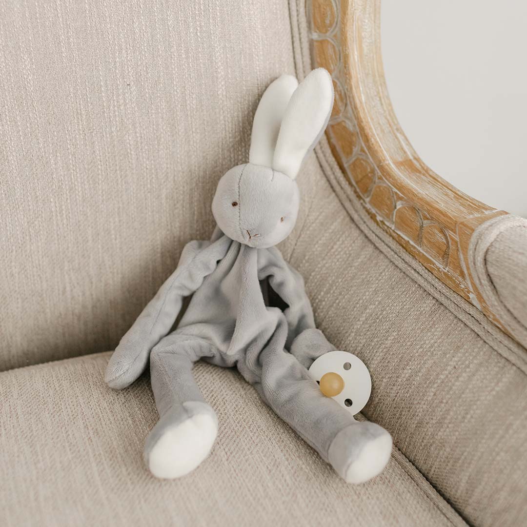 A flat lay photo of the Miles Silly Bunny Pacifier Holder. It is a grey bunny stuffed animal that can hold onto a pacifier