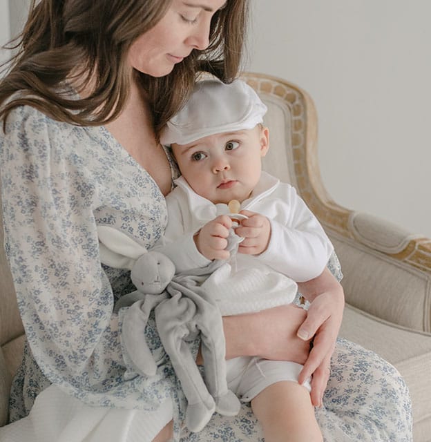 Baby boy with his mother. He is holding onto the Miles Silly Bunny Pacifier Holder, a grey bunny stuffed animal