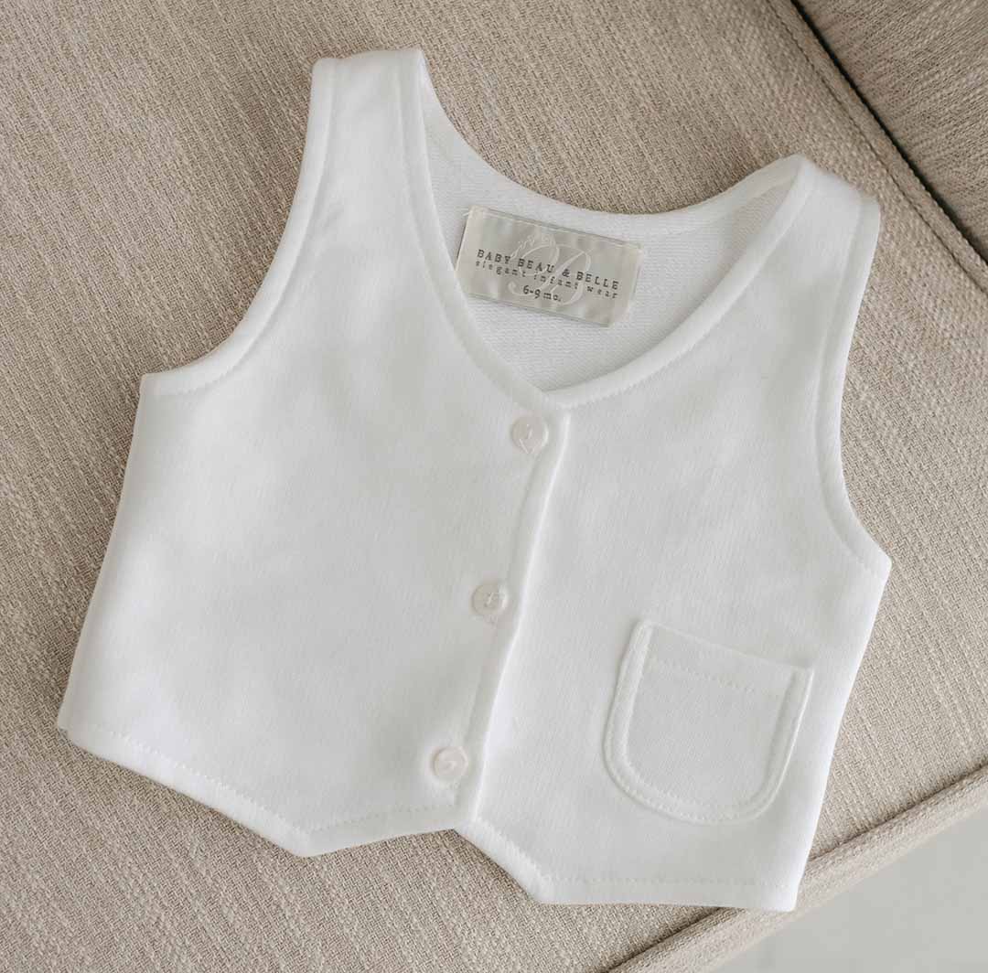The Miles Vest Suit, made from soft French terry cotton in white, includes buttons and a small pocket on the left side. Resting on a beige fabric surface, the interior tag displays "Baby & Child" along with size information. Featuring a V-neck design, it's ideal for pairing with christening booties.