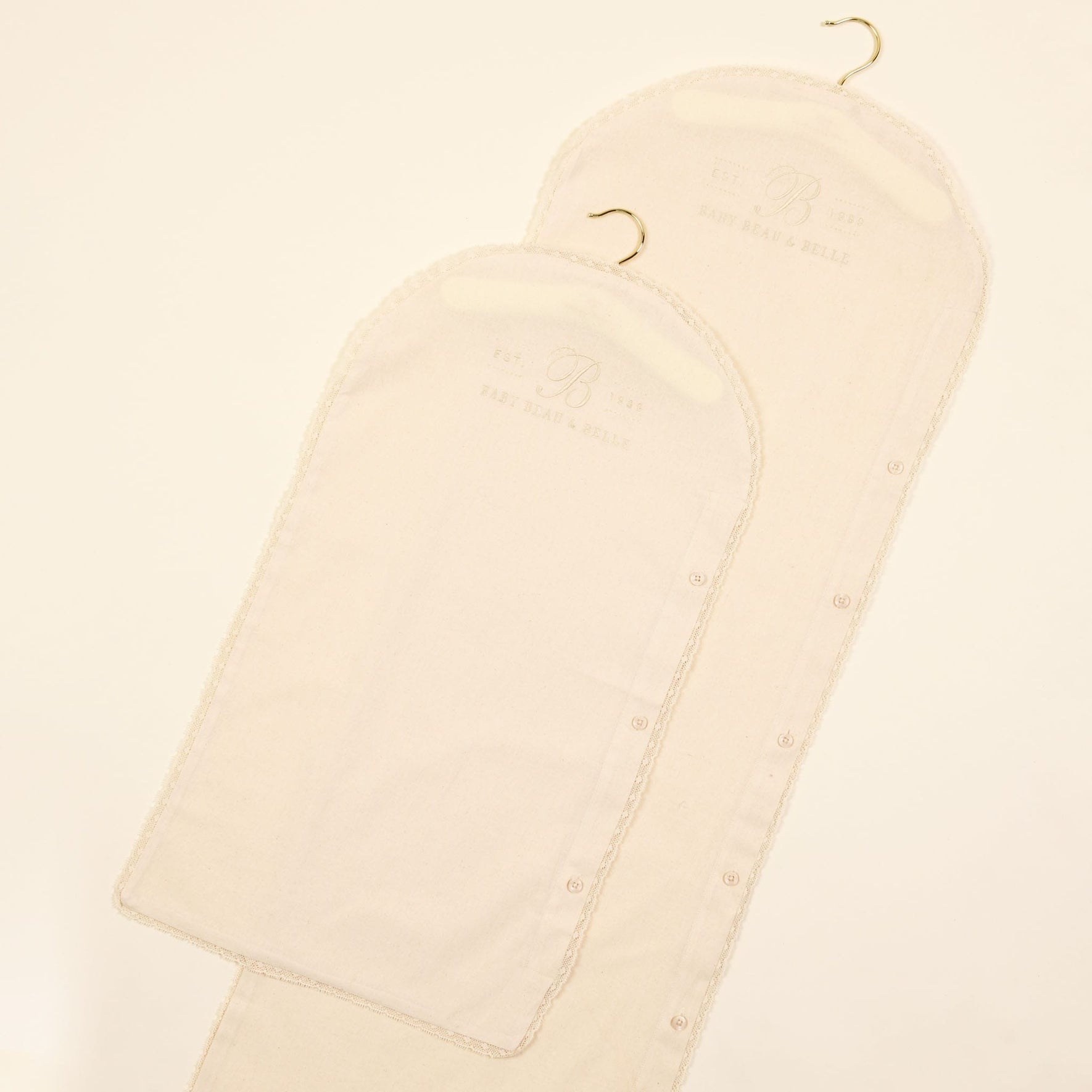 Two Muslin Keepsake Garment Bags in beige, featuring curved tops and small hooks, are laid flat on a white background. These bags have button closures along the sides and subtle embossed text near the top, all crafted from 100% cotton muslin.