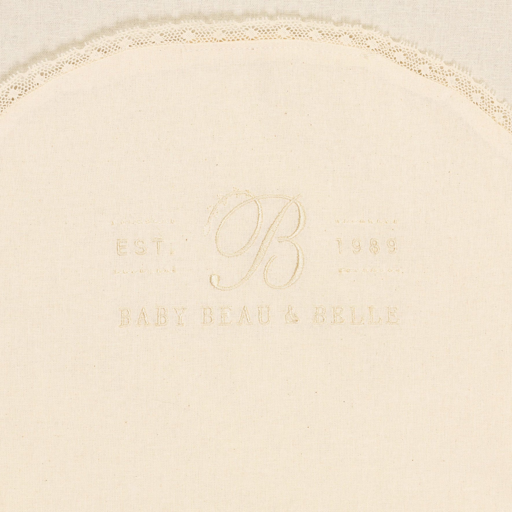 A cream-colored Muslin Keepsake Garment Bag, featuring a delicate lace edging and crafted from 100% cotton muslin. The text "Baby Beau & Belle" is embroidered in the center, along with "Est. 1989" and the letter "B".