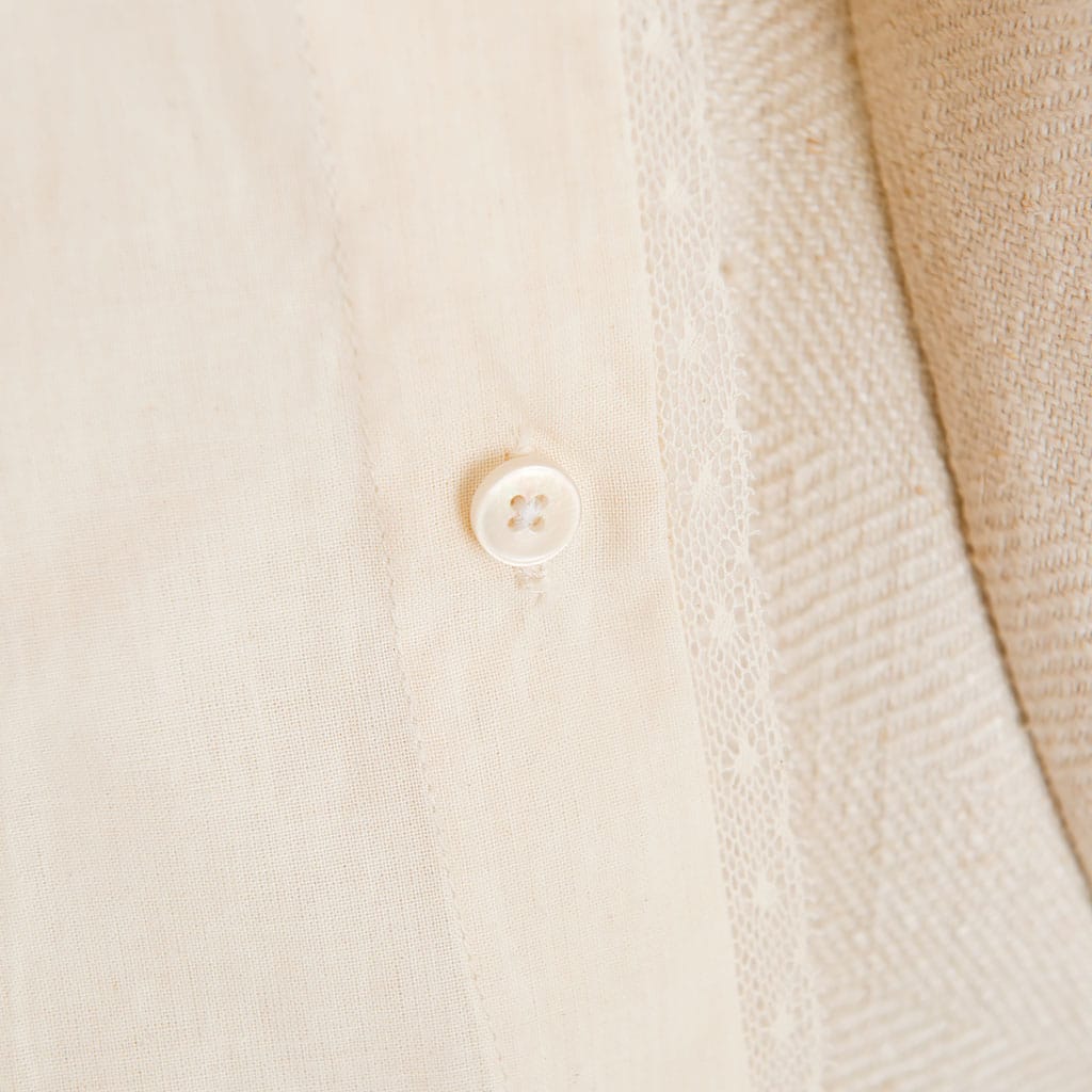 A close-up of a white button on the cream-colored Muslin Keepsake Garment Bag, made from 100% cotton muslin with a subtle pattern. The fabric surrounding the button features stitching details that enhance texture, resembling delicate lace edging. The focus is on the button and the adjacent cloth.