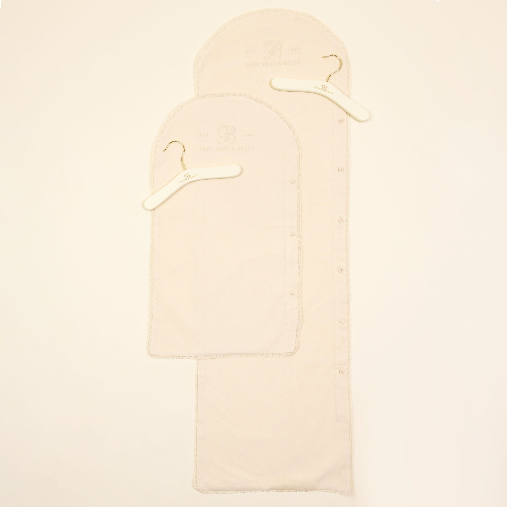 Two Muslin Keepsake Garment Bags of varying sizes are laid flat on a white surface. Crafted from 100% cotton muslin, each bag includes a wooden hanger with a metal hook. Subtle embossed text near the top of the bags adds an elegant detail to their simplicity.