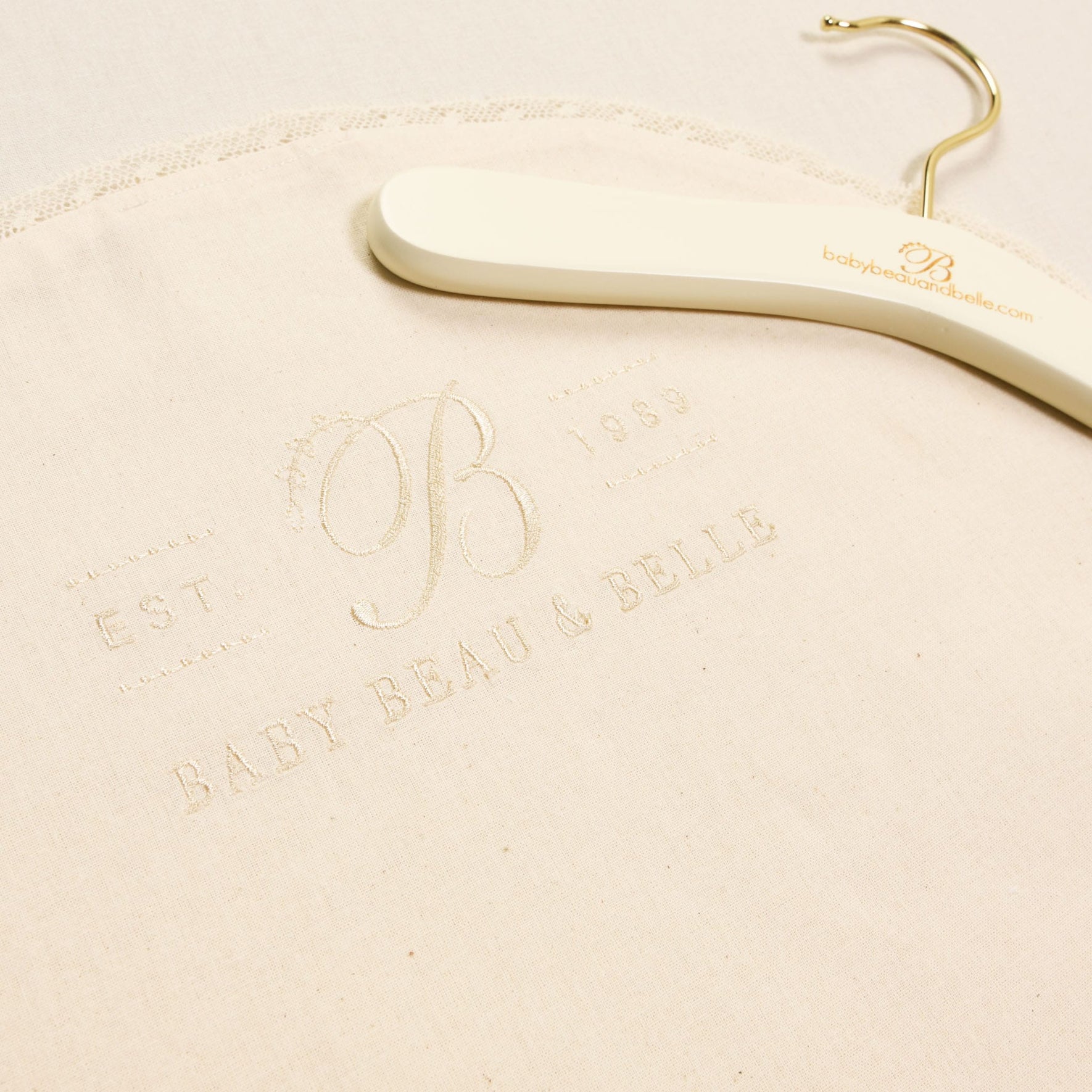 This Muslin Keepsake Garment Bag, crafted from 100% cotton muslin, showcases a subtle embroidery of "Baby Beau & Belle" and "EST 1989" in cream-colored fabric. The elegant design includes a nearby hanger featuring the brand's website, making it perfect for stylish storage.