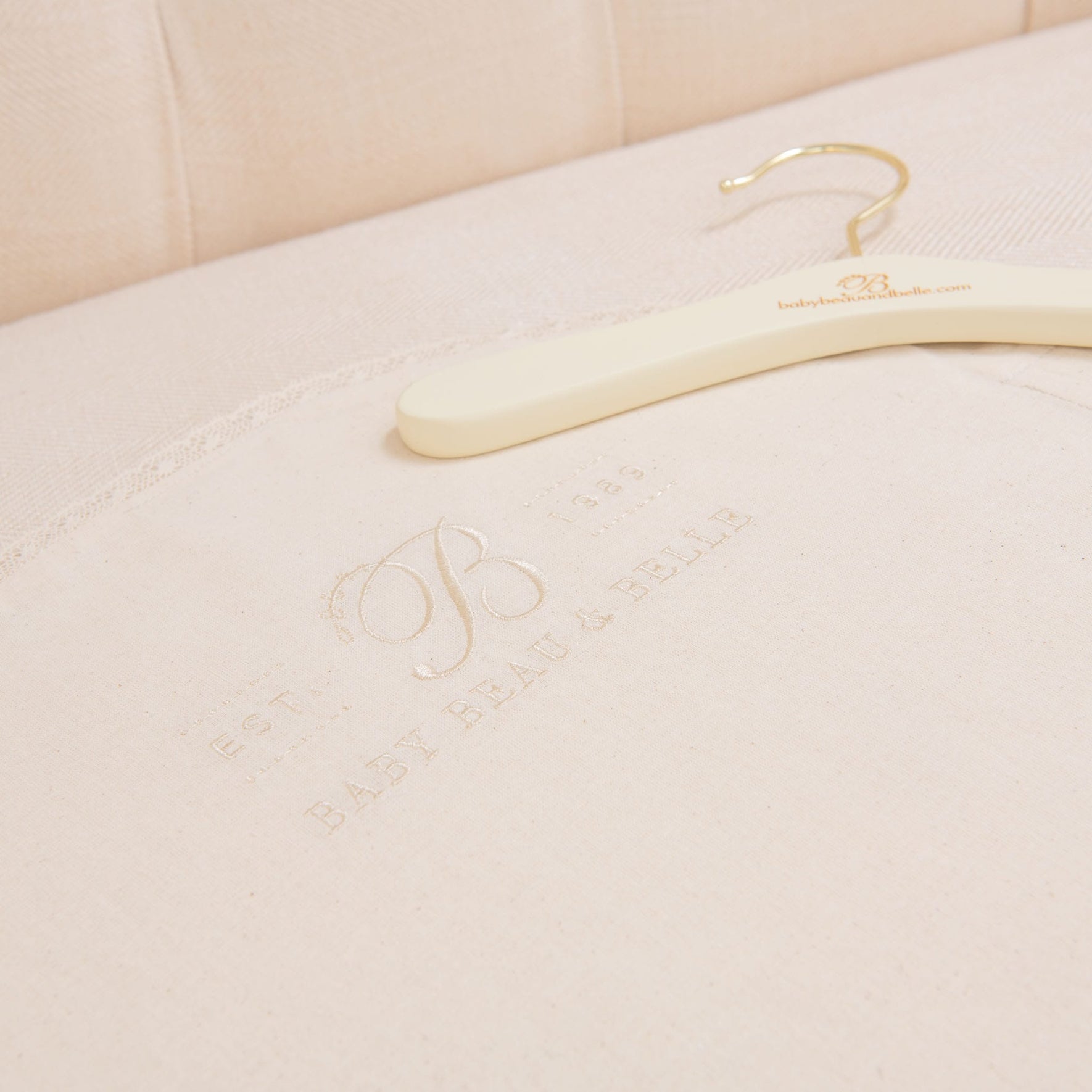 A Muslin Keepsake Garment Bag with a website URL rests on cream-colored fabric, featuring embossed text that reads "EST. 1984 BABY BEAU & BELLE," delicately complemented by lace edging. The cushioned upholstery serves as an elegant backdrop.