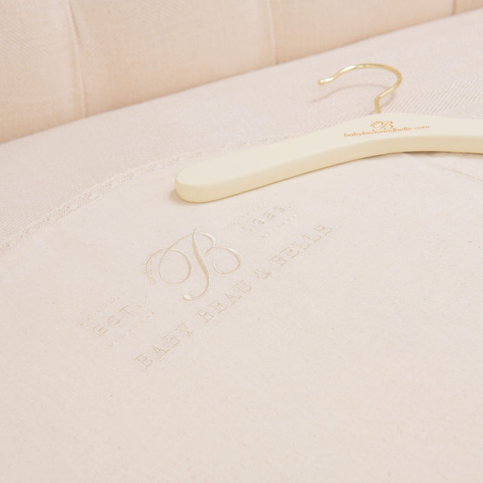 A Muslin Keepsake Garment Bag with a website URL rests on cream-colored fabric, featuring embossed text that reads "EST. 1984 BABY BEAU & BELLE," delicately complemented by lace edging. The cushioned upholstery serves as an elegant backdrop.