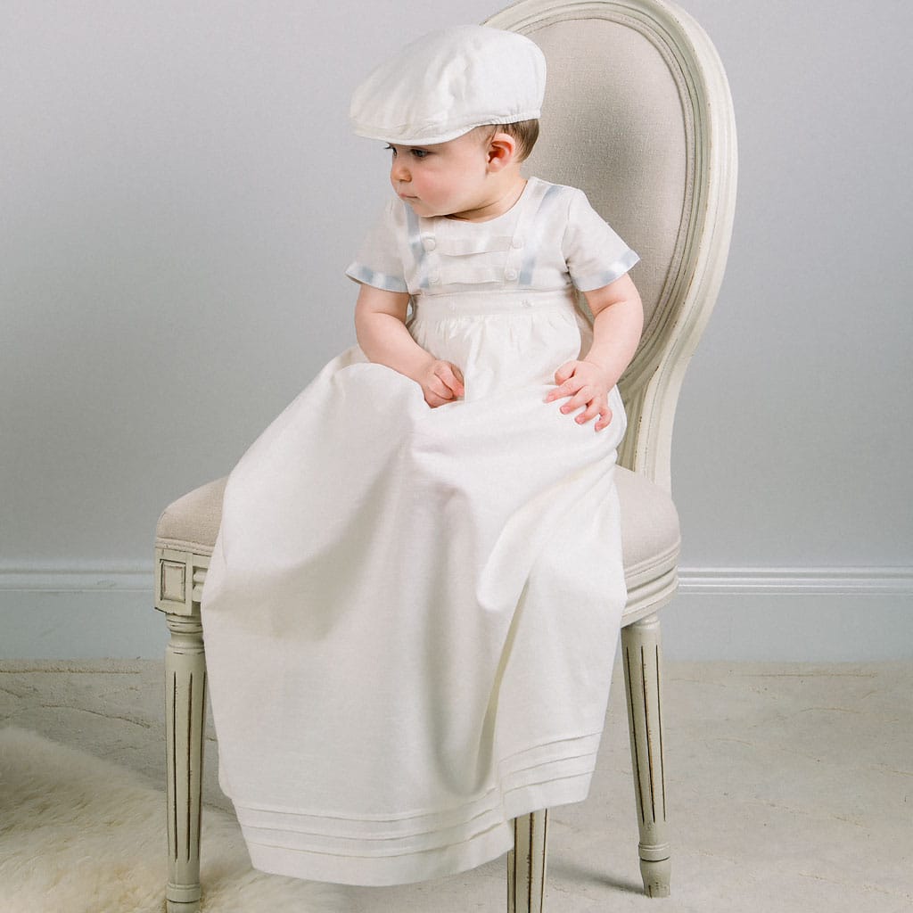 A young child sits on a white upholstered chair, adorned in the Owen Linen Convertible Christening Skirt with a matching cap, looking to the side. The light gray wall and beige floor subtly highlight the elegance of their attire.