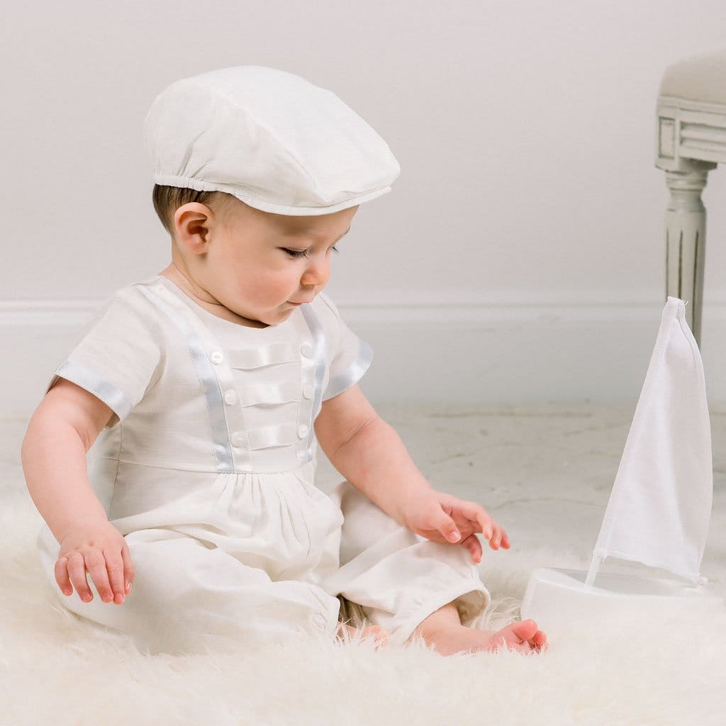 Dressed in the Owen Romper from the Accessory Bundle, a baby sits on a fluffy surface, gazing down at a tiny white sailboat toy. The minimalistic background's neutral color palette perfectly complements the scene.
