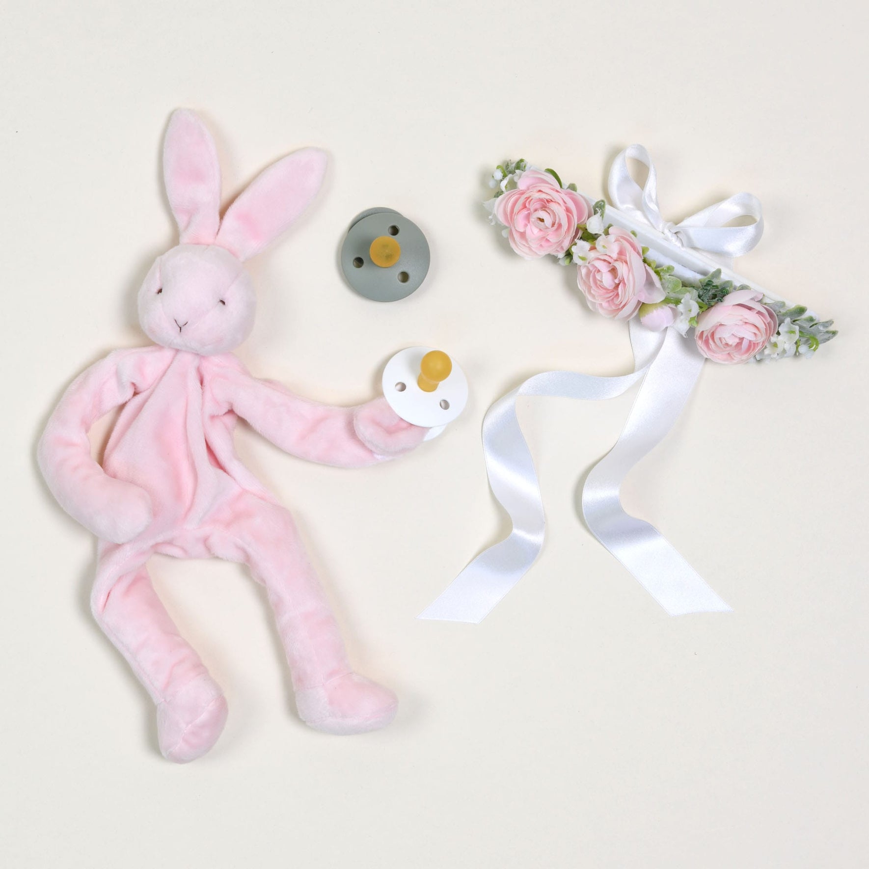 The Pink Silly Bunny Buddy & Pacifier features a soft, plush pink bunny that cuddles a white pacifier, complemented by a gray pacifier clip that's positioned nearby. The ensemble includes a decorative floral headband embellished with pink flowers and white ribbons, all elegantly arranged on a light background for baby-safe enjoyment.