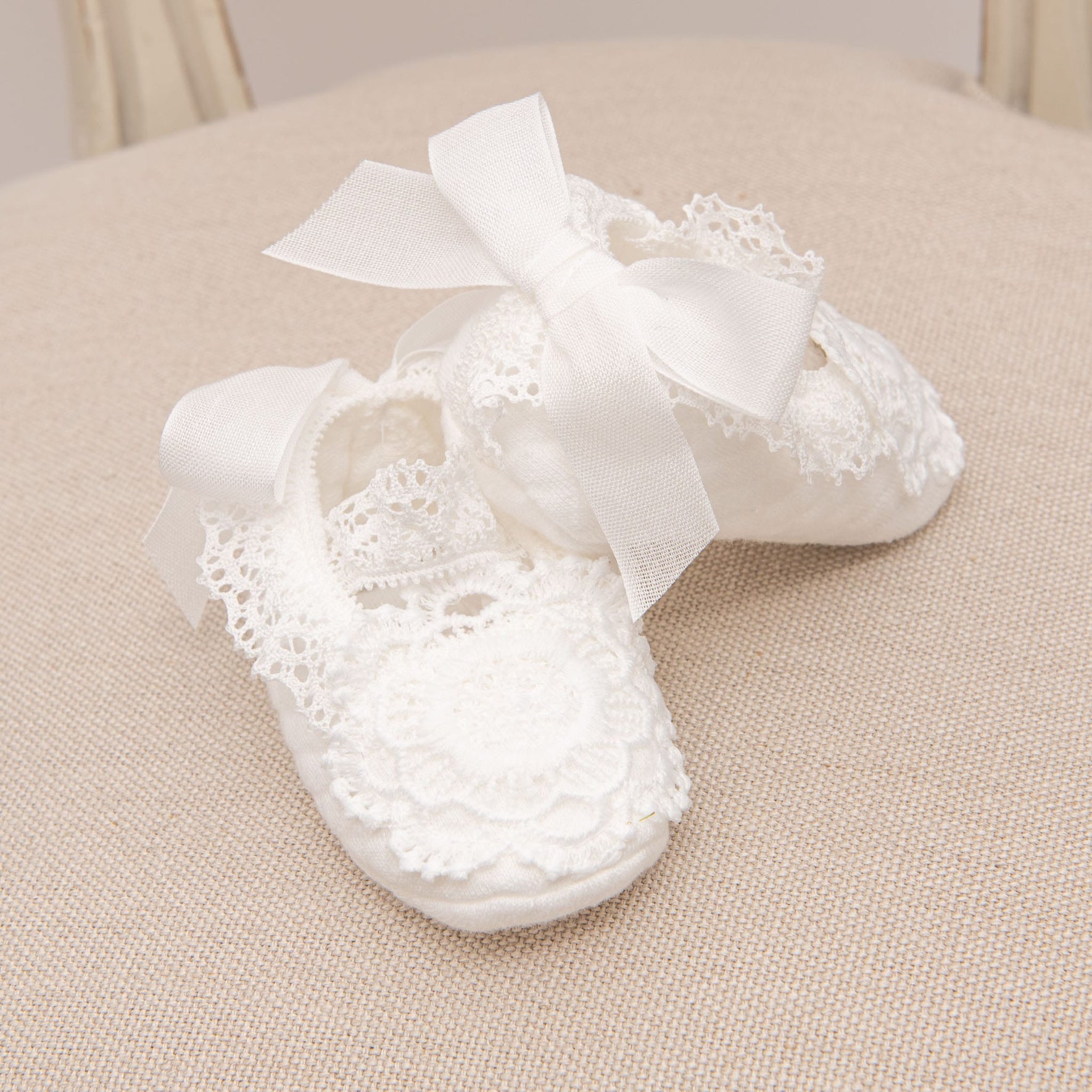 Poppy Christening Booties with intricate floral lace and satin ribbon ties, arranged neatly on a beige fabric surface.