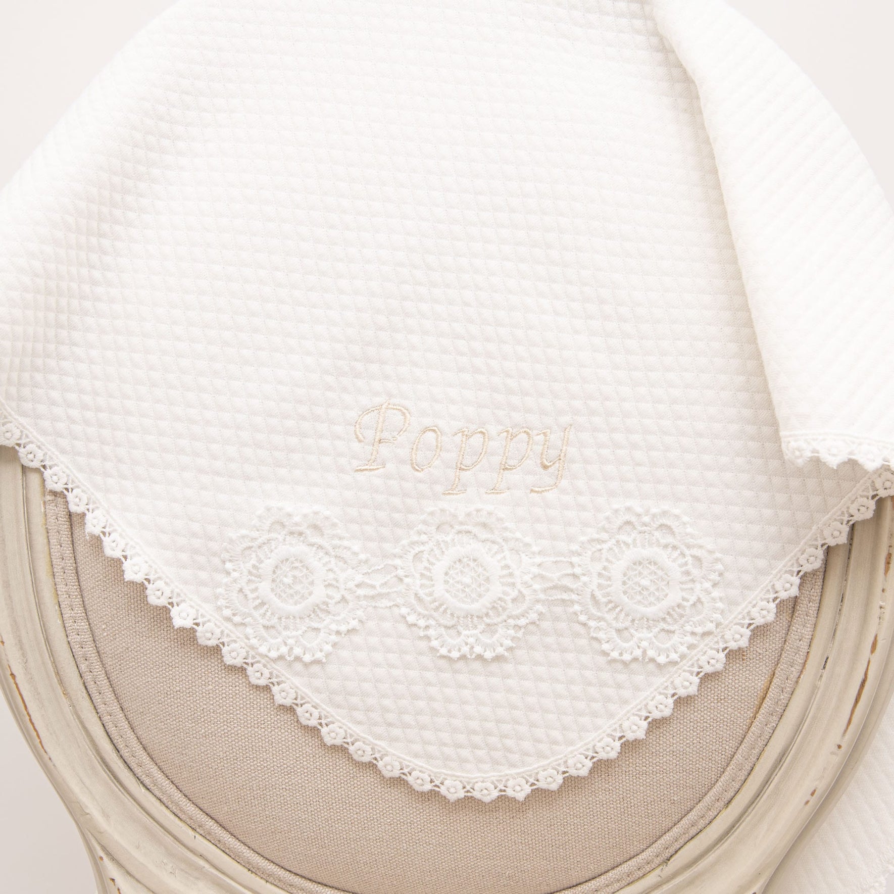Quilted Poppy Christening Blanket elegantly displayed on a chair, showcasing delicate floral lace trim and 'Poppy' embroidery.