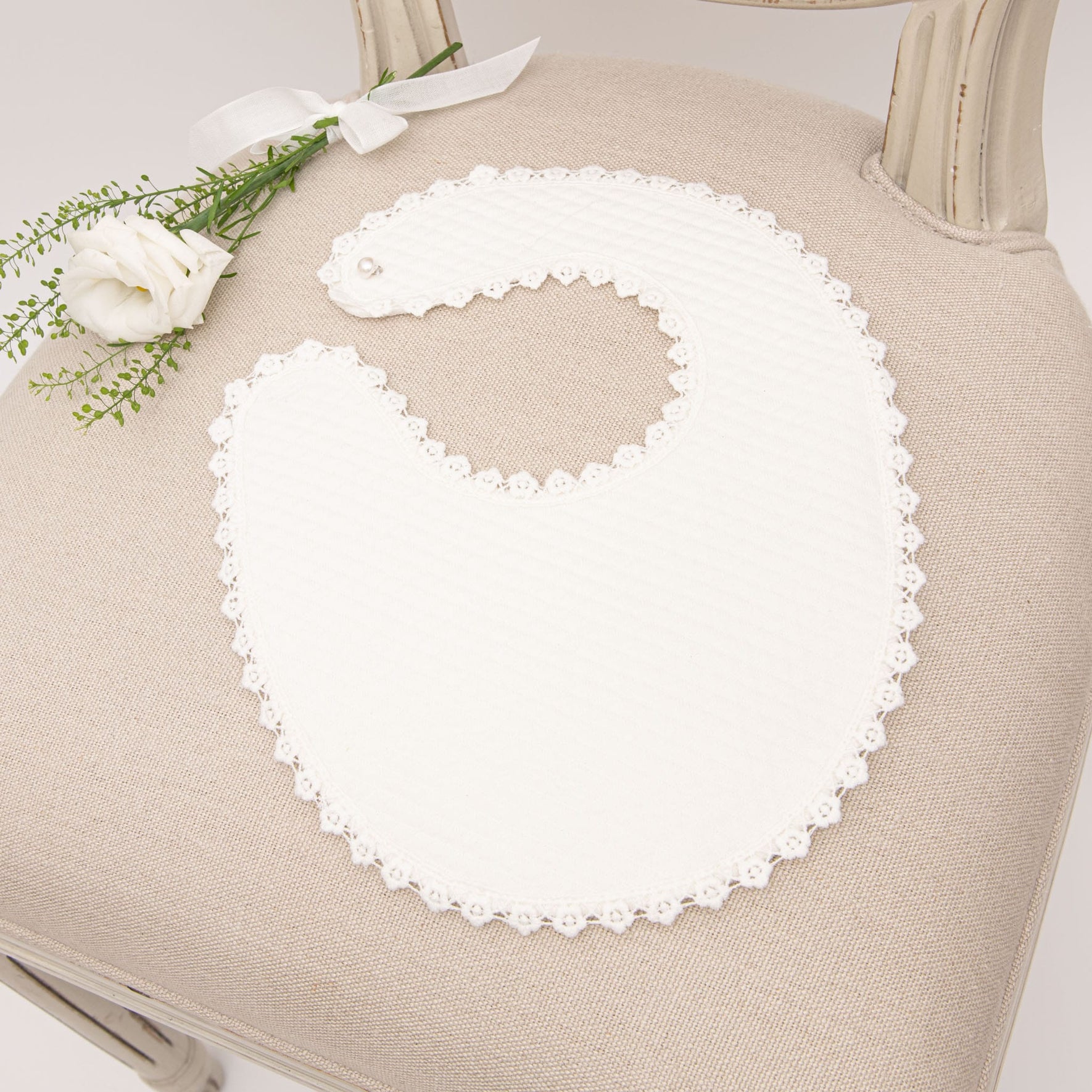 The Poppy Christening Bib laid on a neutral fabric chair, showcasing soft quilted cotton and elegant scalloped lace detailing, accented with a white flower arrangement.