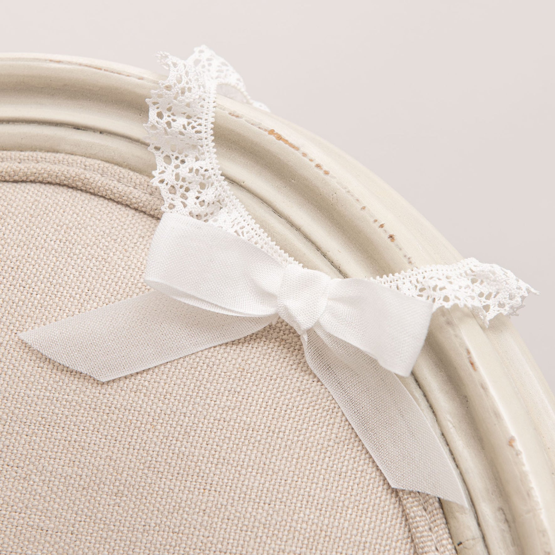 Detailed view of the Poppy Christening Headband, showcasing a white bow and lace accents, placed on a vintage chair.