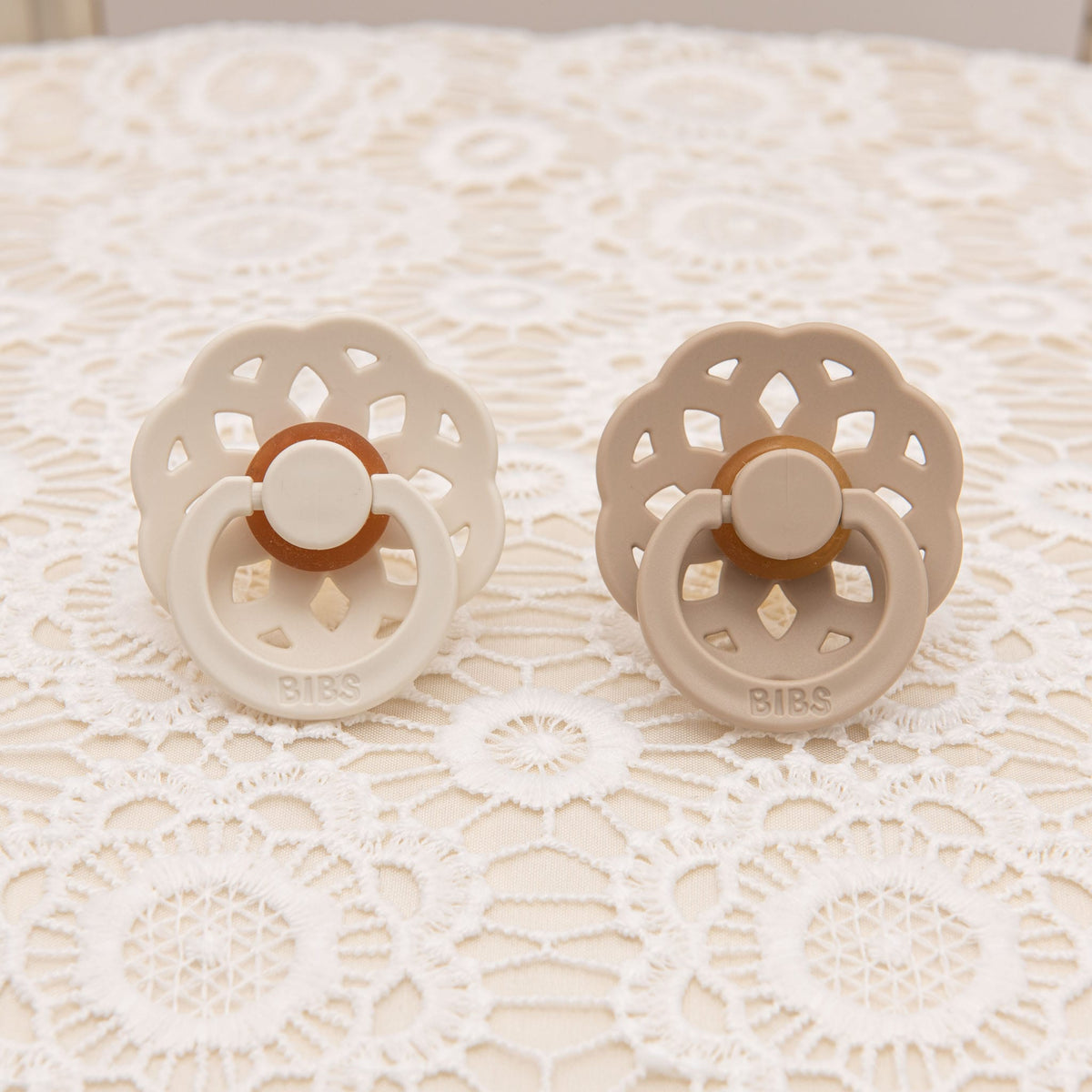 Vanilla and ivory pacifier set with scalloped edges, styled on intricate floral lace for a charming presentation.