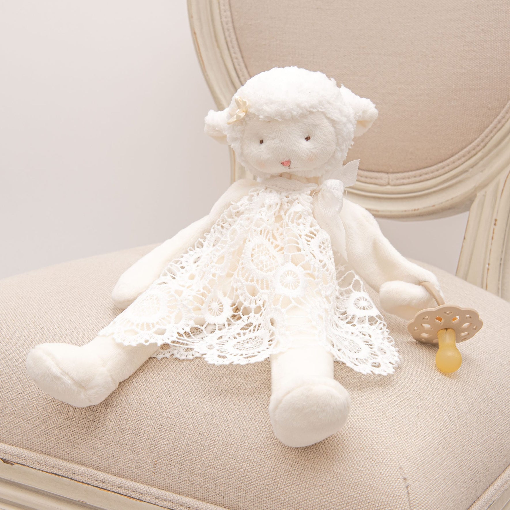 The Poppy Silly Lamb Buddy Pacifier Holder displayed on a beige chair, a soft lamb adorned with lace details coordinating with the Poppy Christening Gown, holding a pacifier.