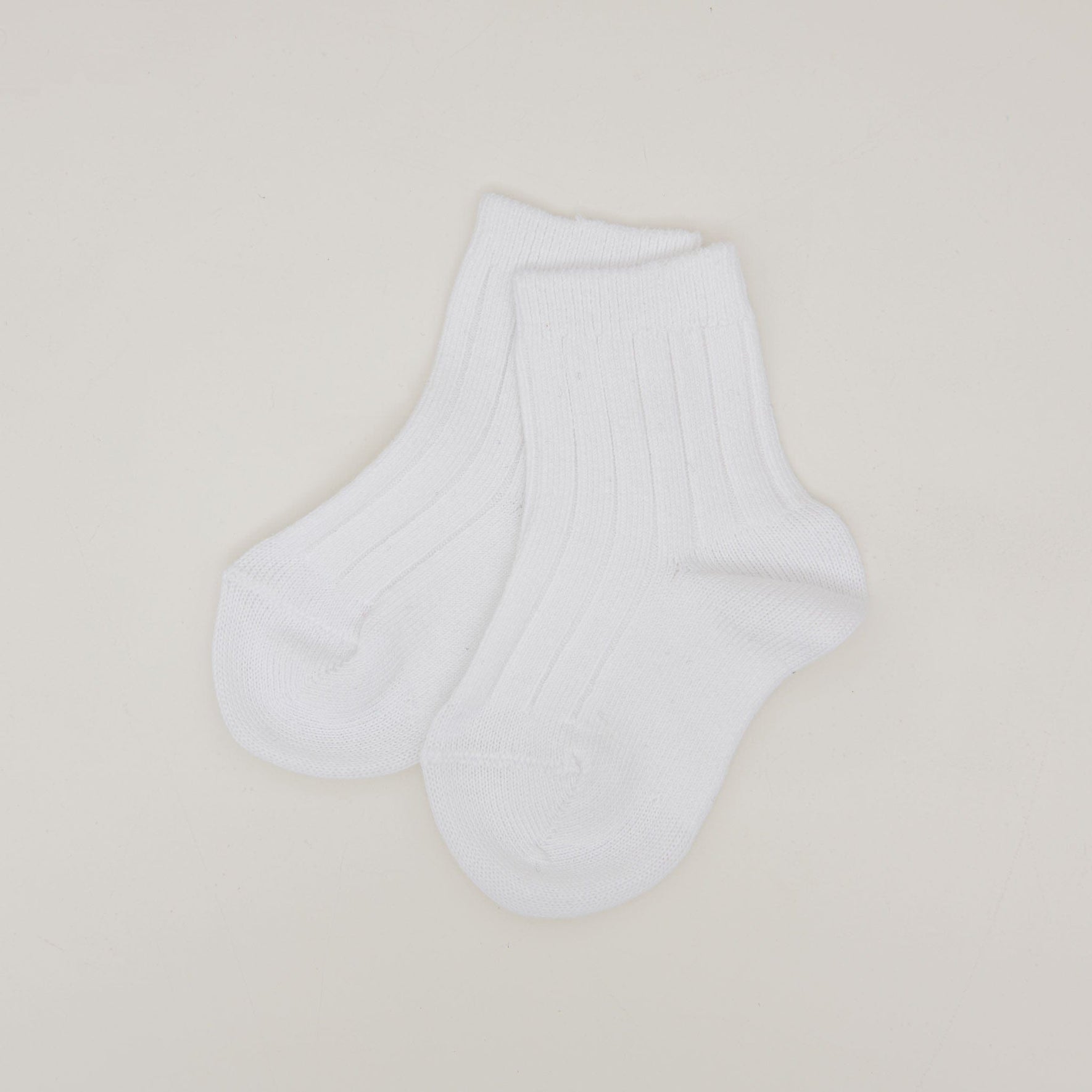 A pair of Ribbed Socks, crafted from a white cotton blend and made in Spain, is displayed flat against a simple light background. The socks have a soft appearance with distinct ribbed detailing, and the toes and heels are designed with a subtle rounded shape.