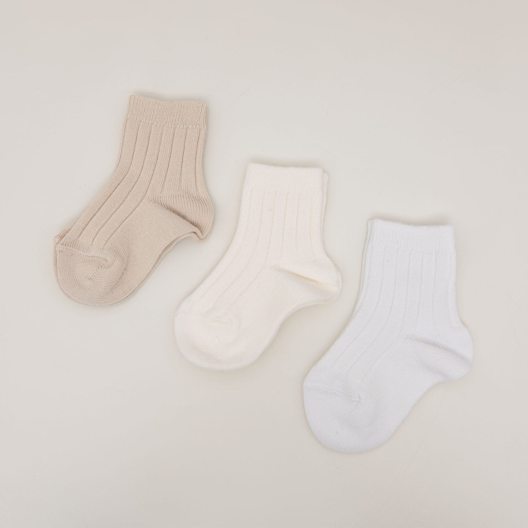 Three pairs of Ribbed Socks, made from a gentle cotton blend, are showcased on a neutral backdrop. Crafted in Spain, these cozy socks are laid out side by side in calming shades of beige, cream, and white.